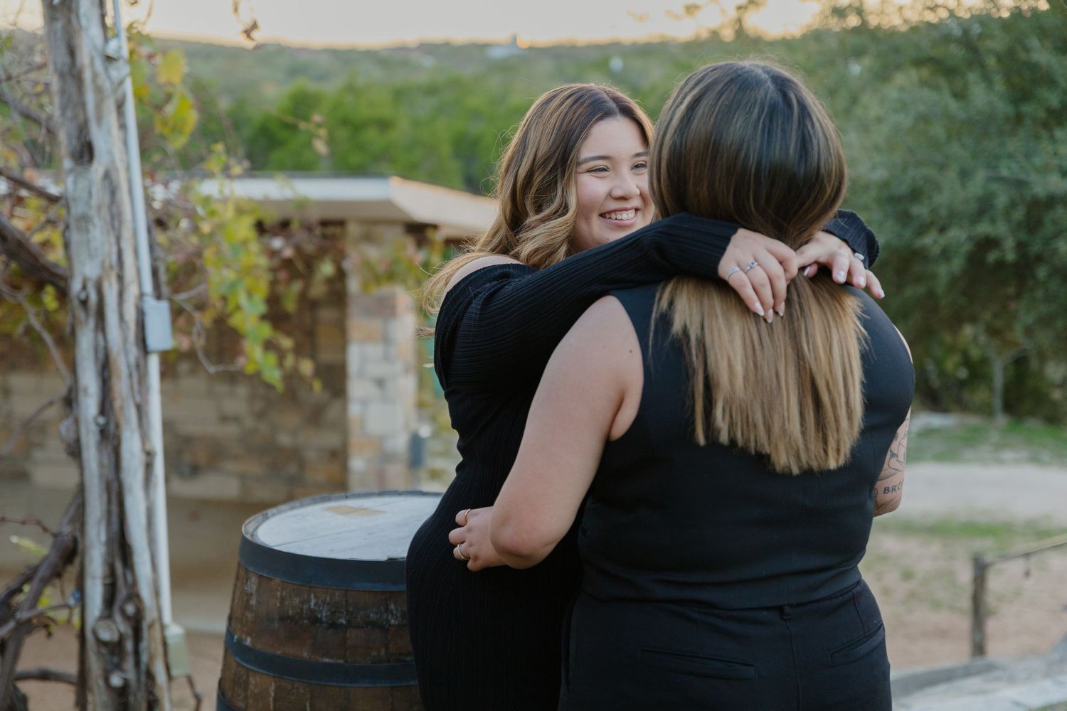Friends share emotional embrace near rustic wine barrel in outdoor setting.