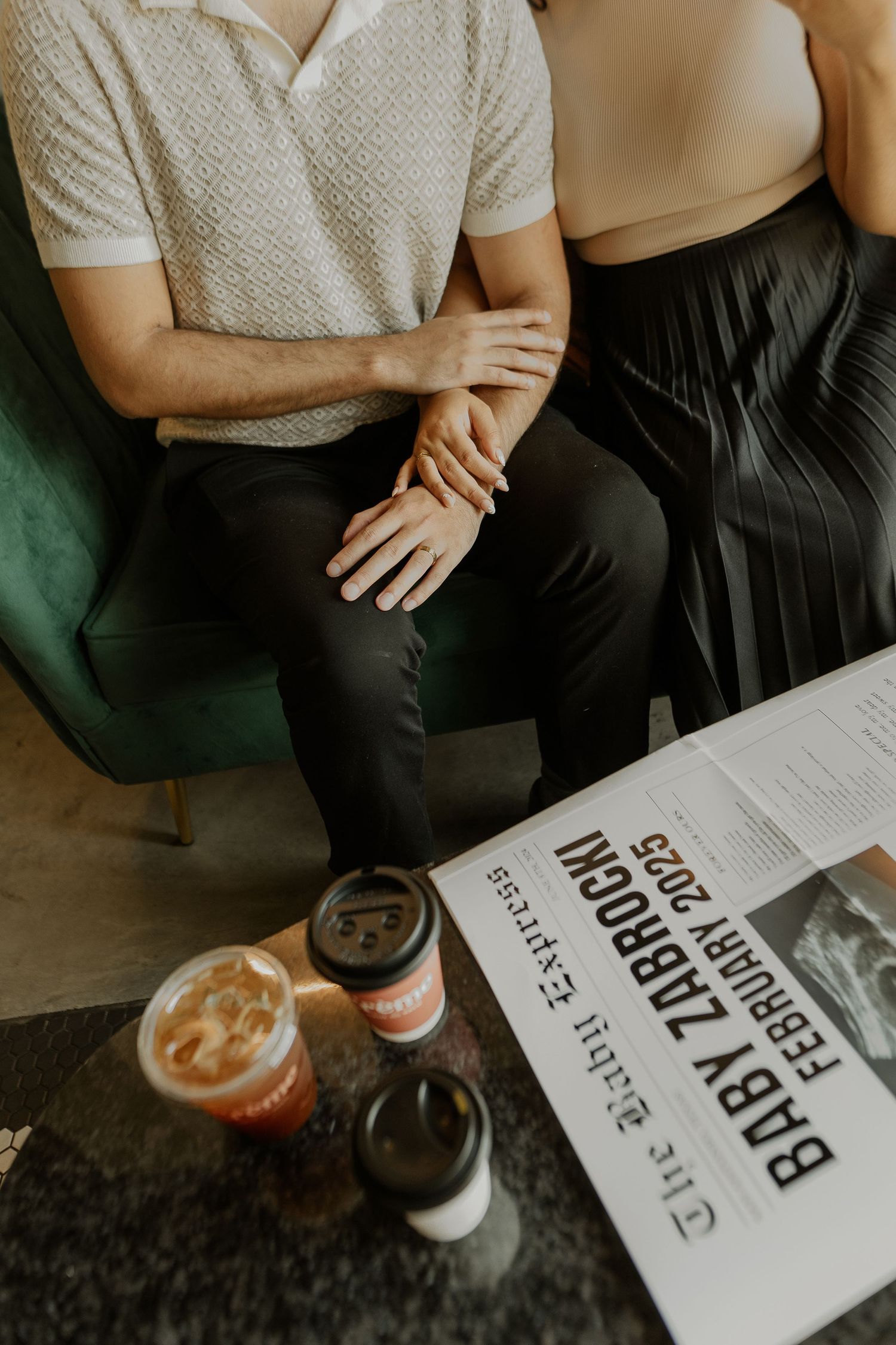 Couple sits on green couch with coffee drinks and newspaper on table in cozy indoor setting.