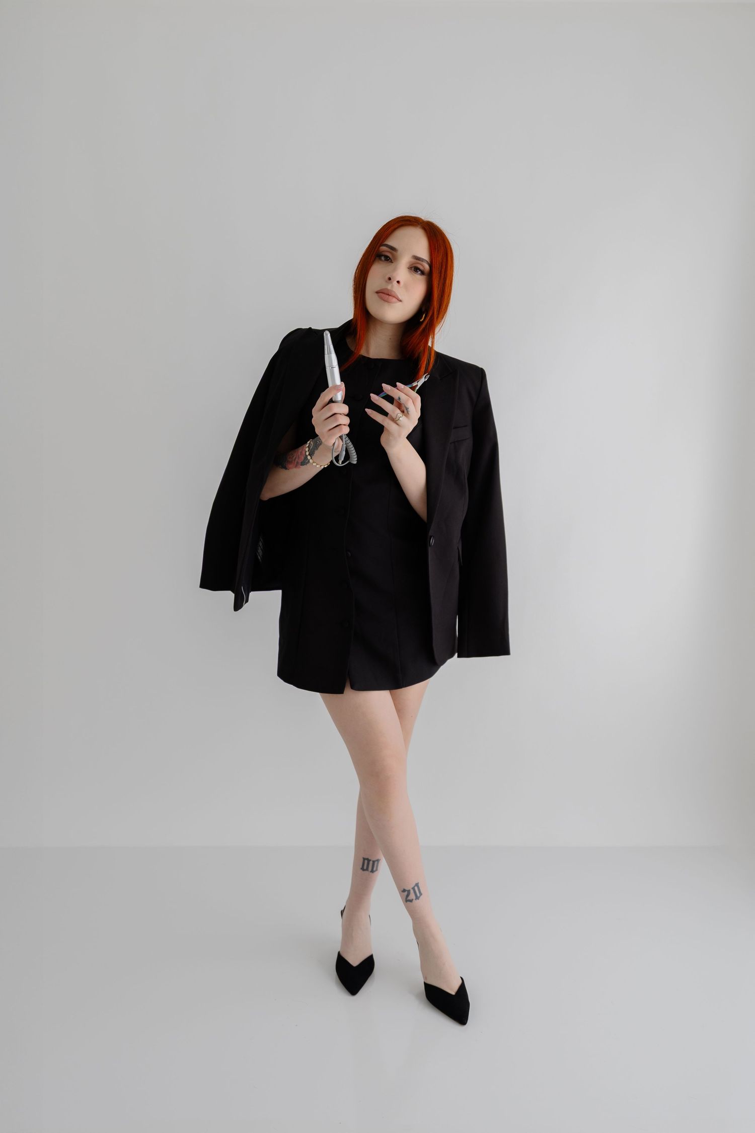 A person in a stylish black blazer and dress poses against a white backdrop with red hair and heeled shoes.