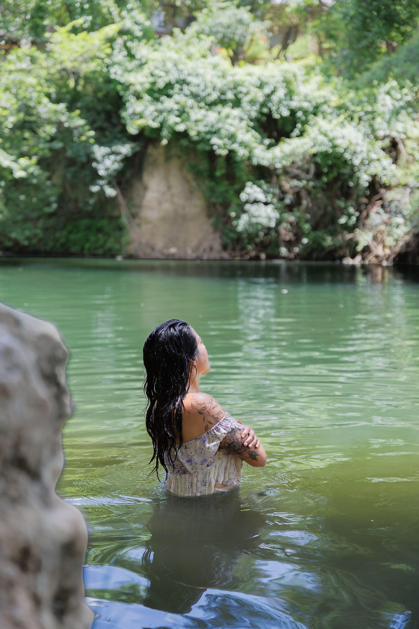 Woman with wet dark hair standing waist-deep in a serene green river surrounded by lush trees and white flowers.