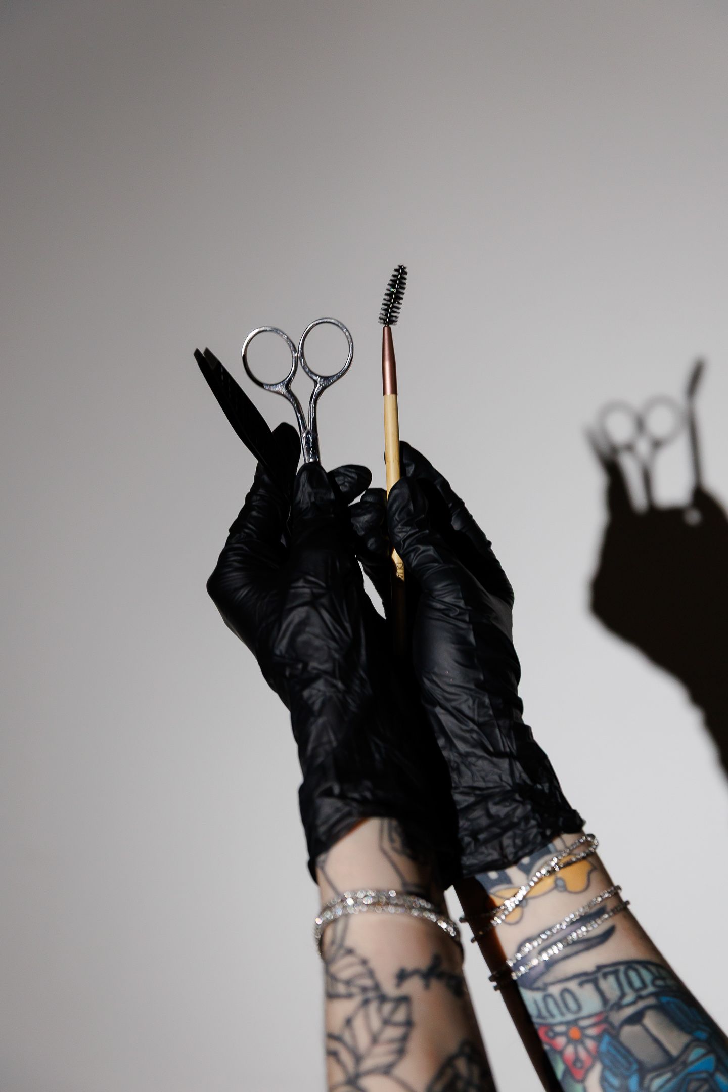 Tattooed hands in black latex gloves holding eyebrow scissors and spoolie brush against gray background.