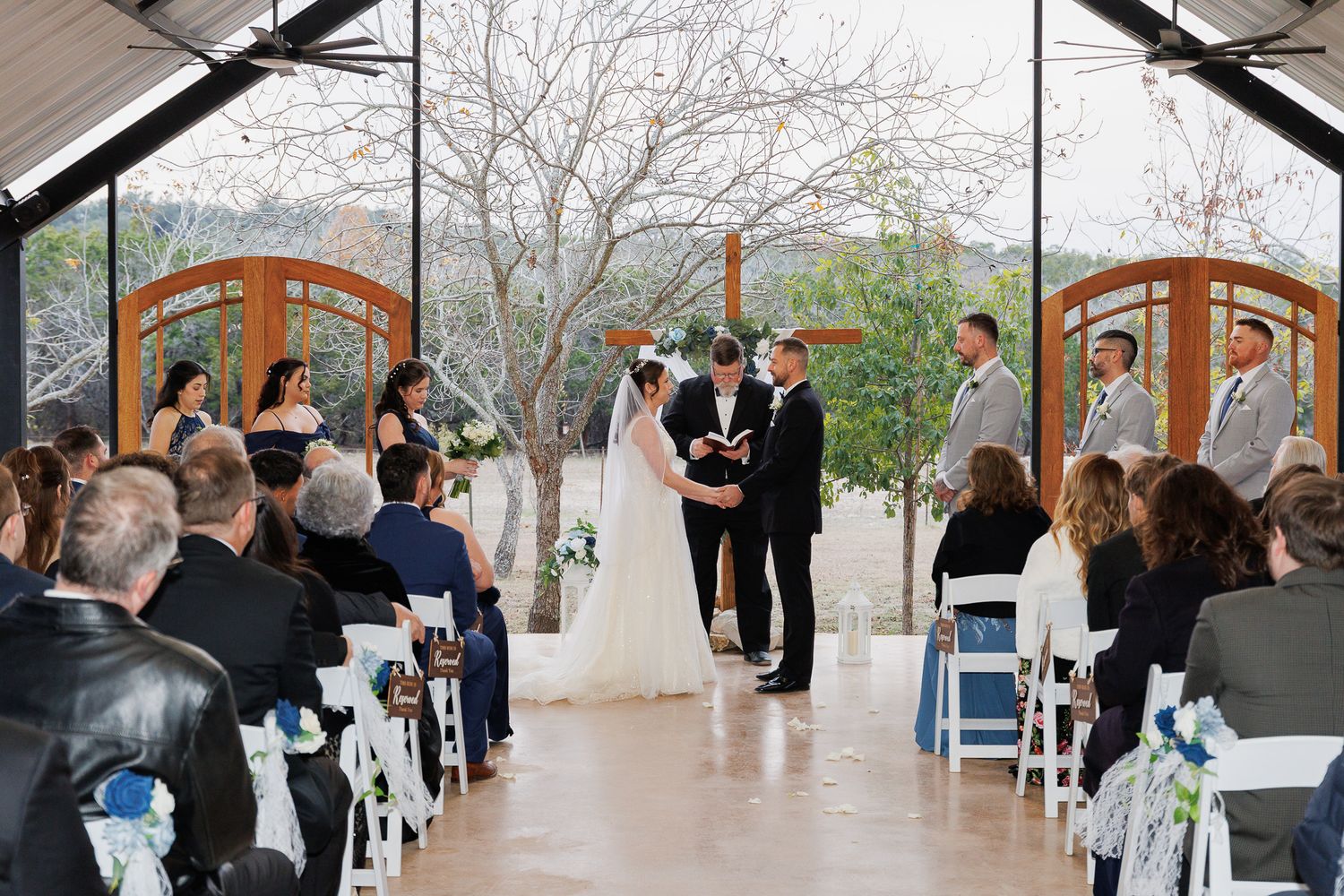 Wedding ceremony guests gather inside an elegant rustic venue with wooden beams and large windows.