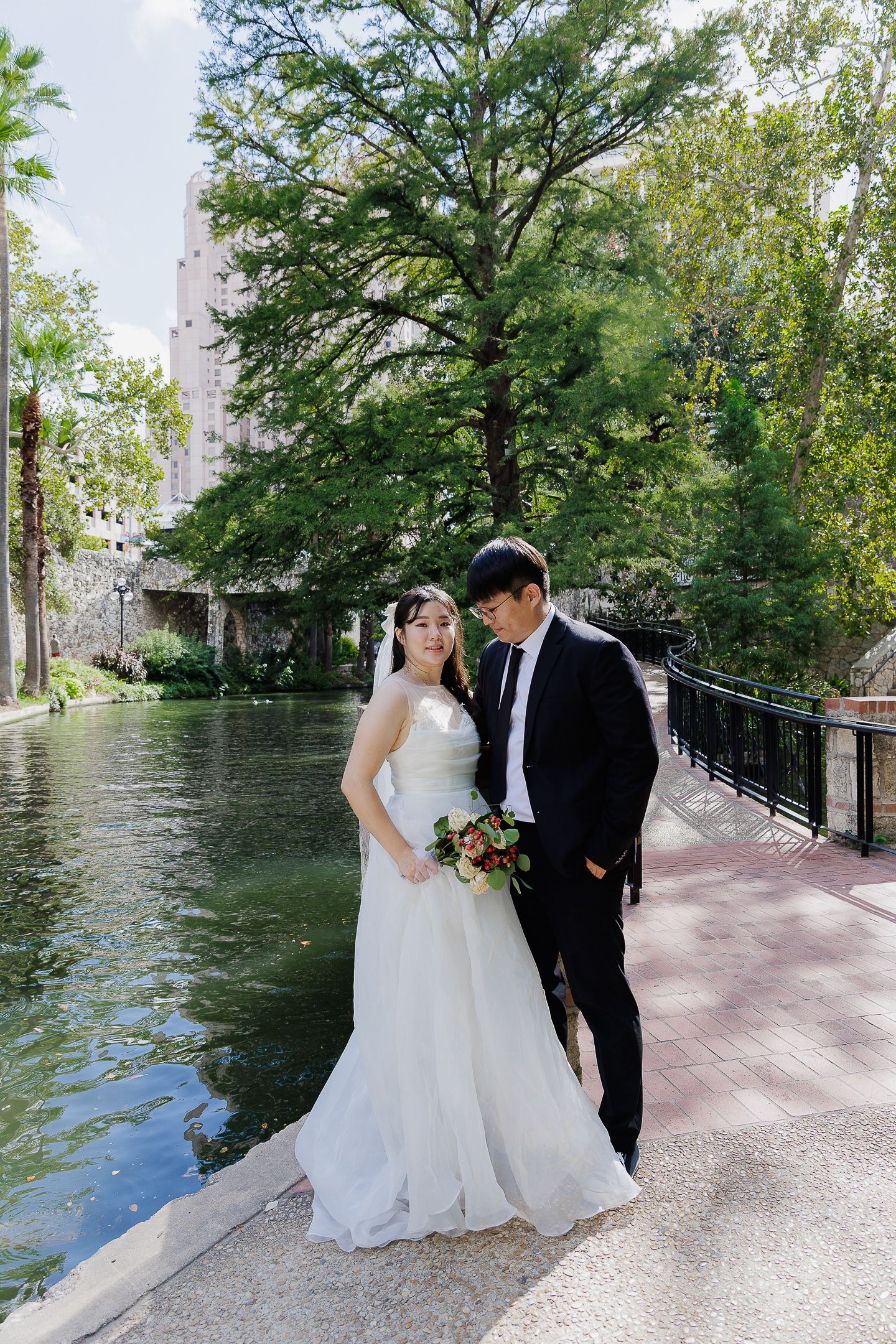 A couple shares a romantic moment along a waterfront walkway with lush green trees and a tranquil river in the background.