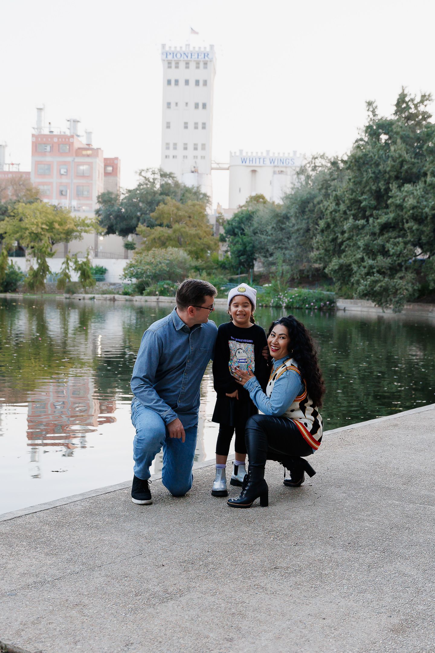 A couple sits together by a tranquil pond with city buildings visible in the background.
