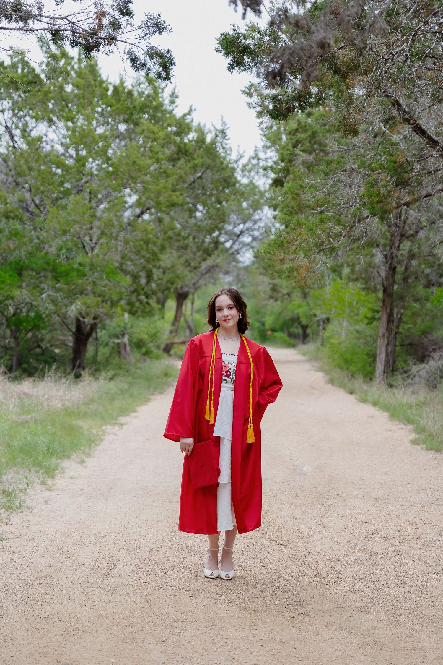 Graduate wearing traditional red graduation gown standing on dirt path surrounded by trees.