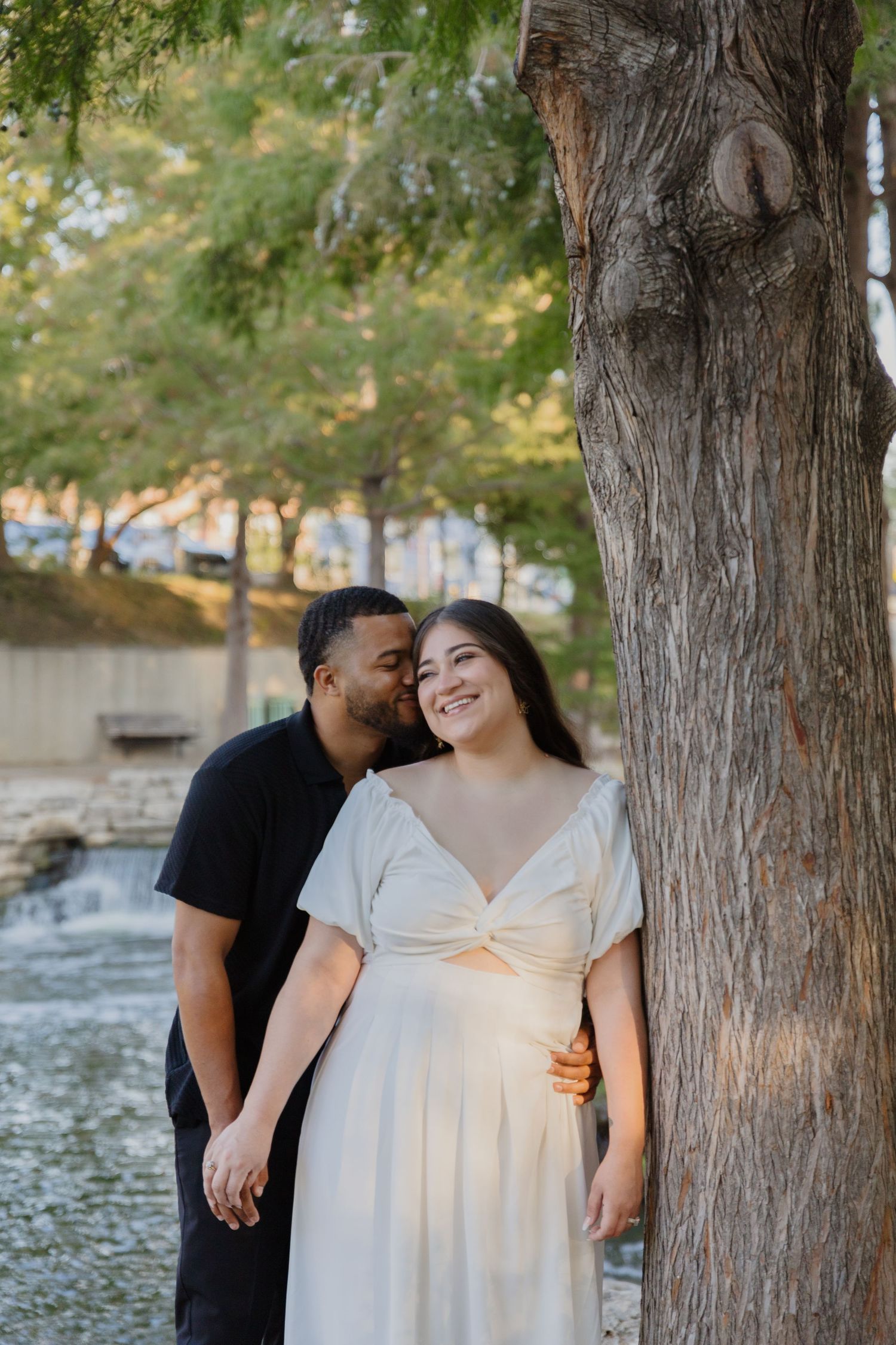 A couple shares a tender moment by a tree near a flowing water feature.