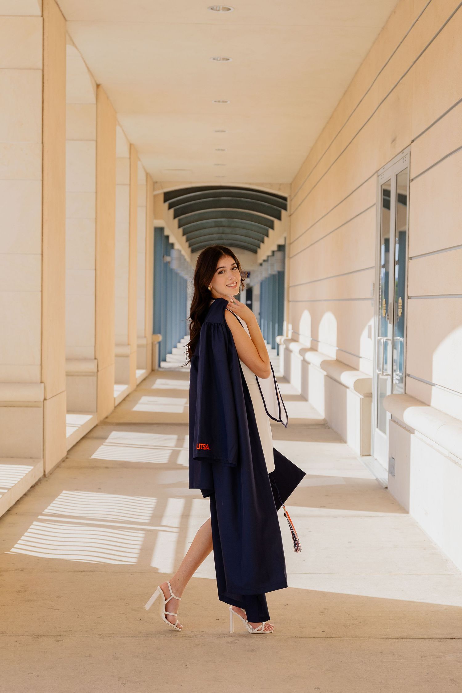 Graduate in navy cap and gown poses in sunlit campus hallway with architectural columns.