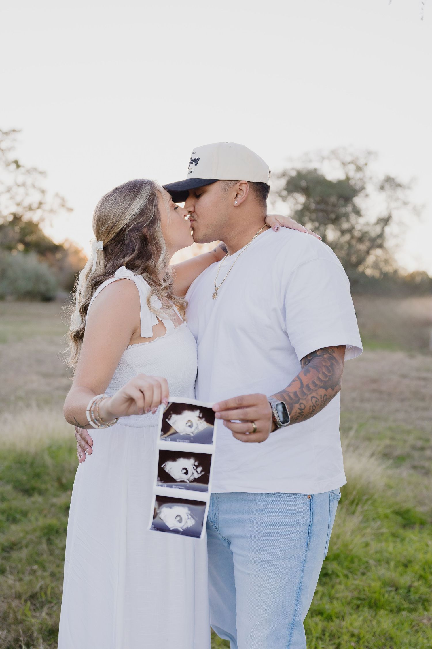 Couple sharing an intimate moment while holding ultrasound photos in an outdoor setting.