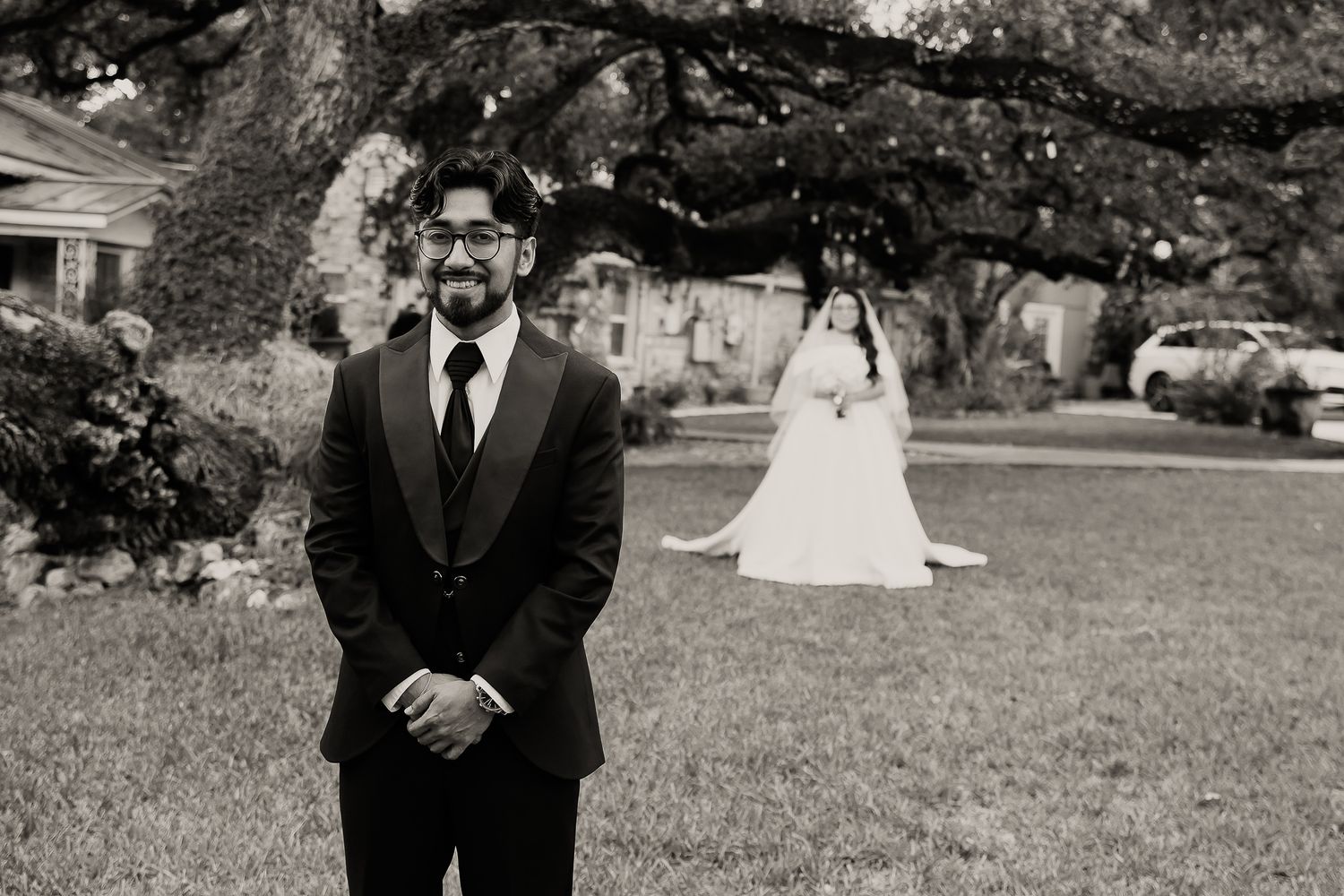 A black and white wedding photo shows people standing outdoors with trees and greenery in the background.