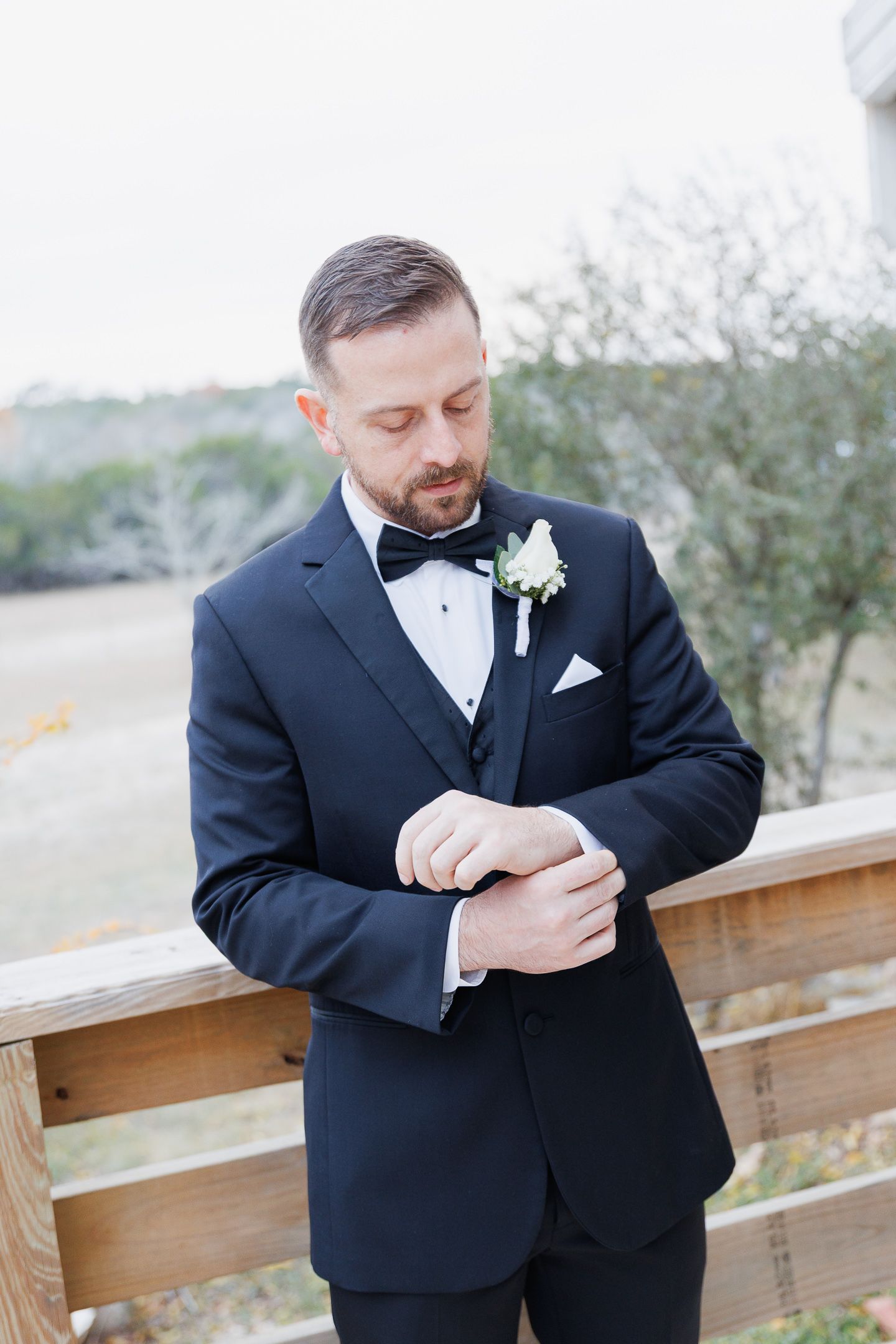 A person in a black tuxedo adjusts their cufflinks while standing against a wooden railing outdoors.
