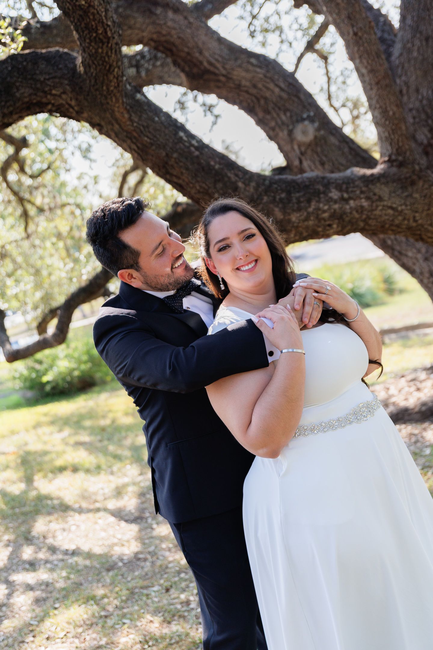 A playful wedding photoshoot outdoors under large oak trees showing joyful moments between newlyweds.