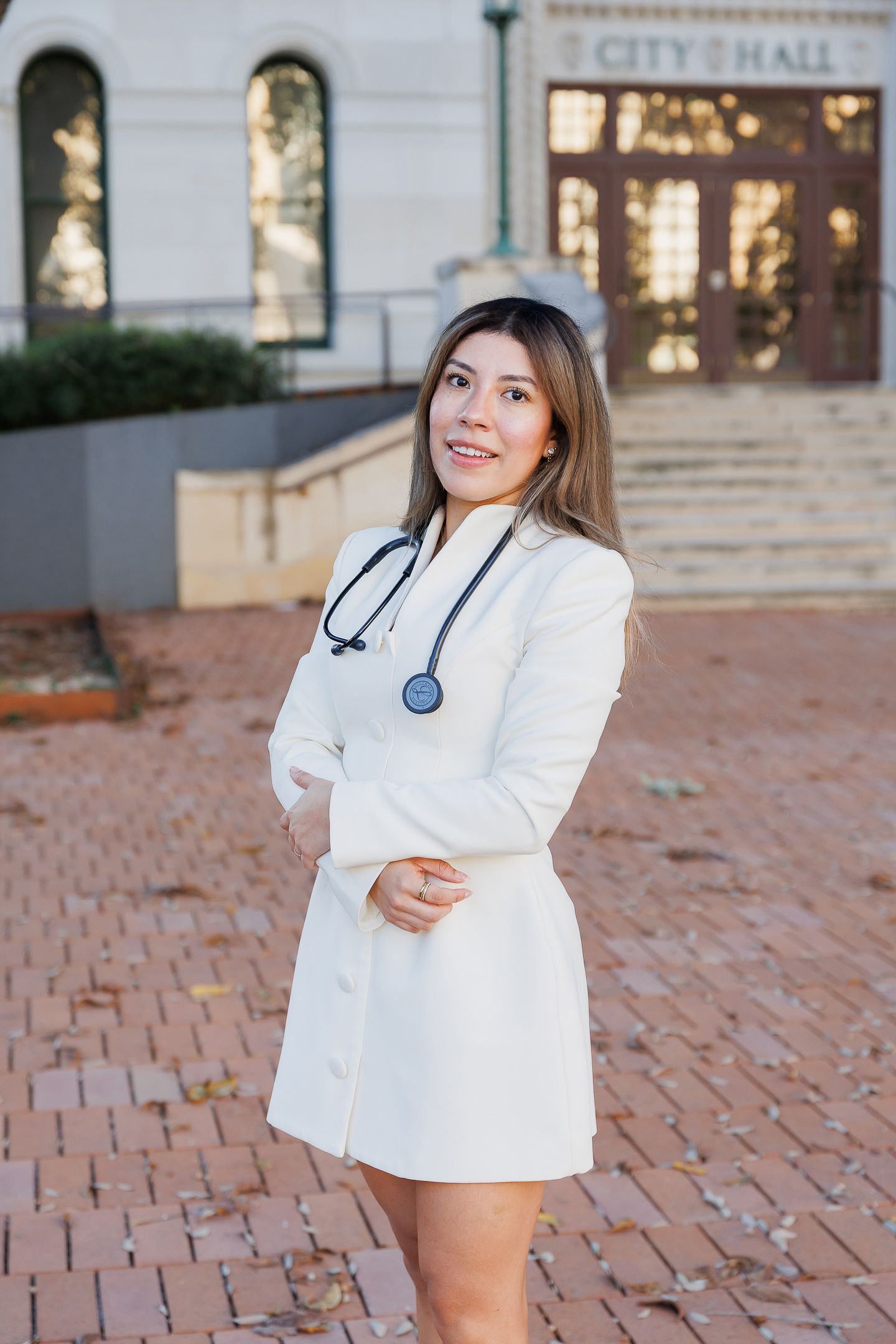 A medical professional in a white coat and stethoscope poses in front of a brick building with City Hall signage.