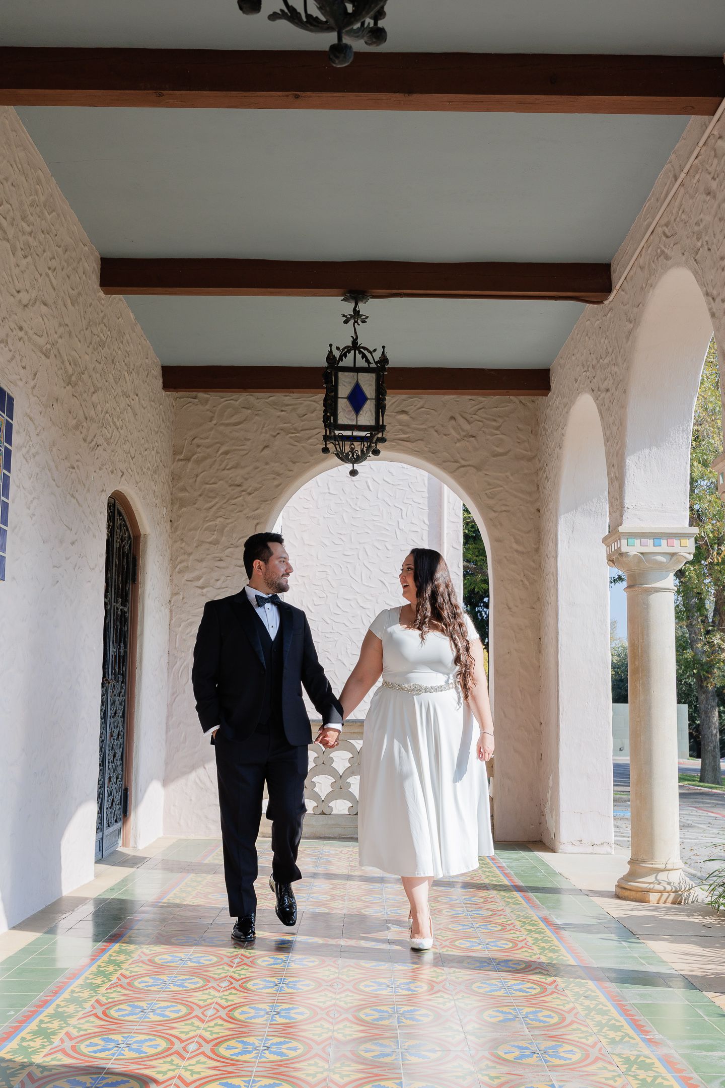 Couple walks together through Spanish-style archway with ornate tiled floors and white stucco walls.