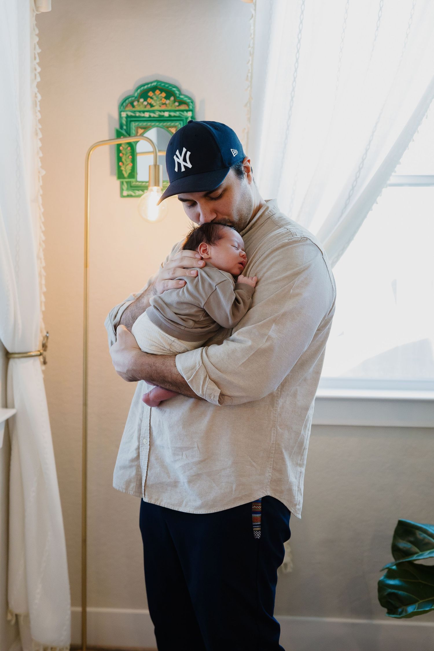 A tender moment between an adult wearing a Yankees cap and a sleeping baby in a light-filled room with green wall decor.