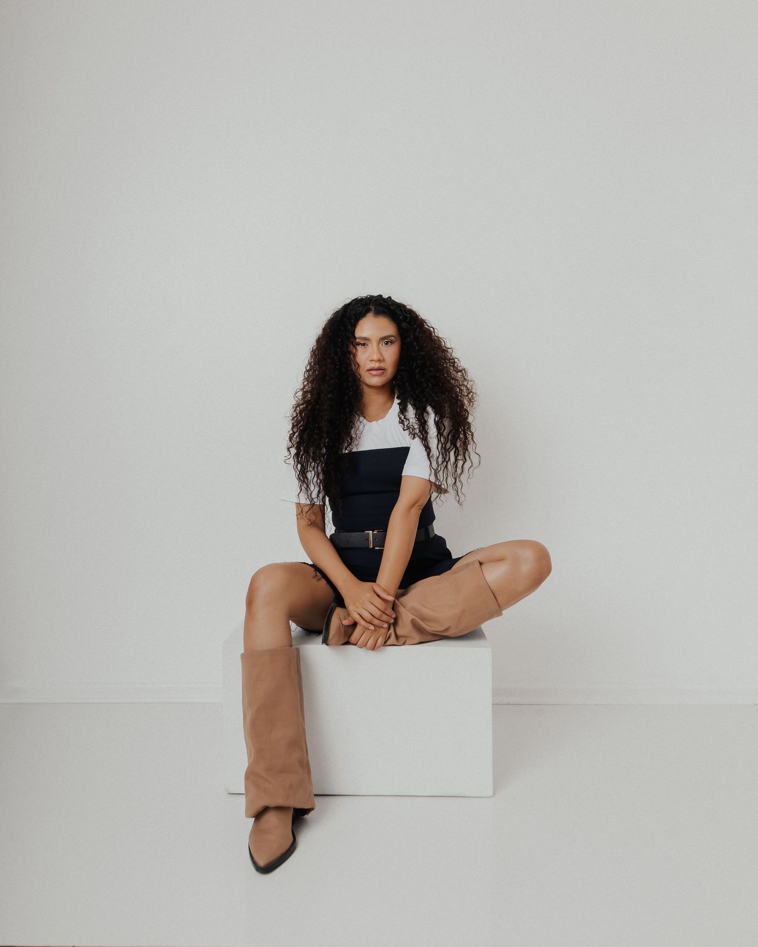 A model in casual attire sits confidently on a white cube against a neutral background.