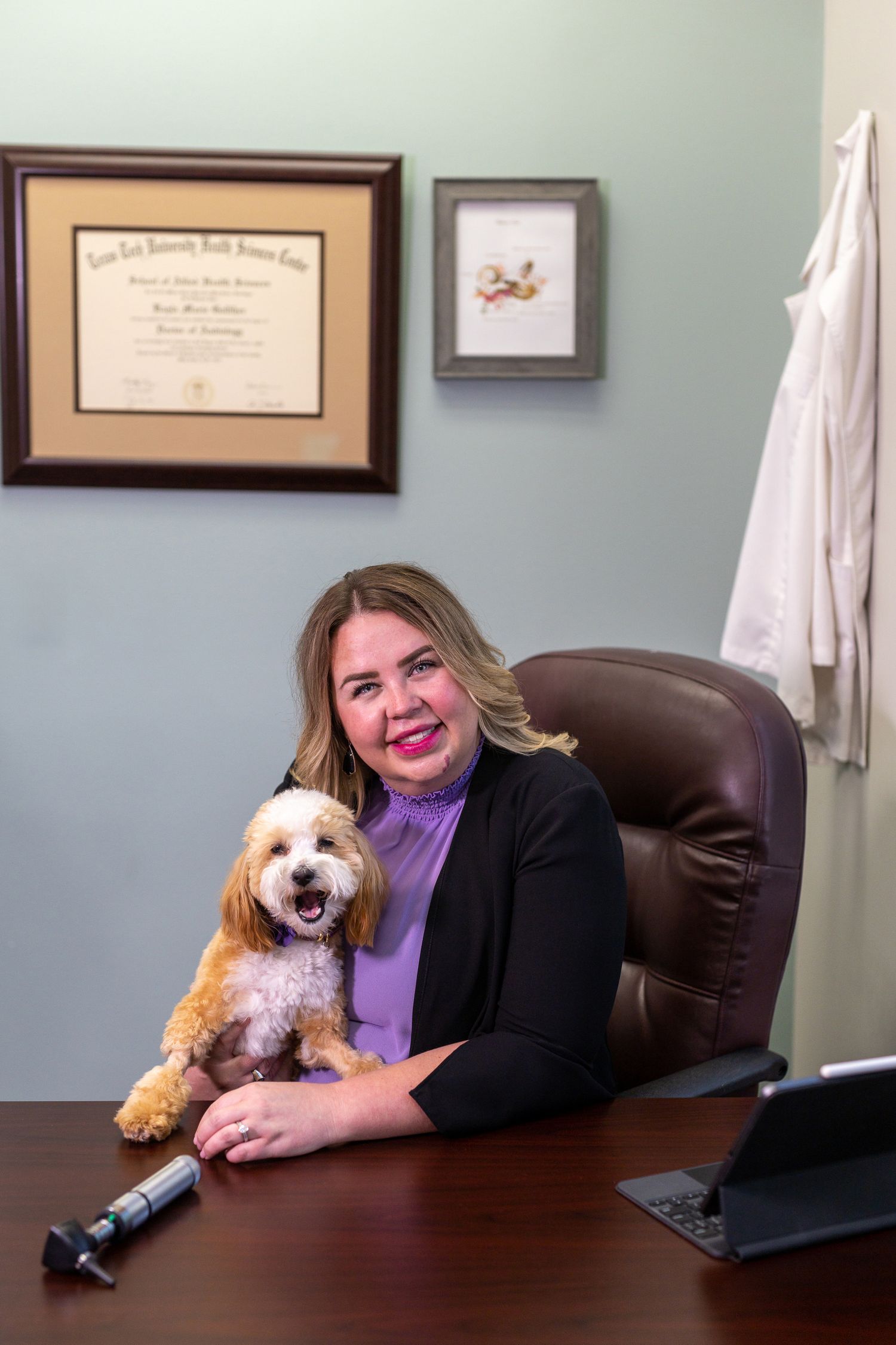 Professional woman in purple top and black blazer sits at desk with small fluffy dog in medical office setting.