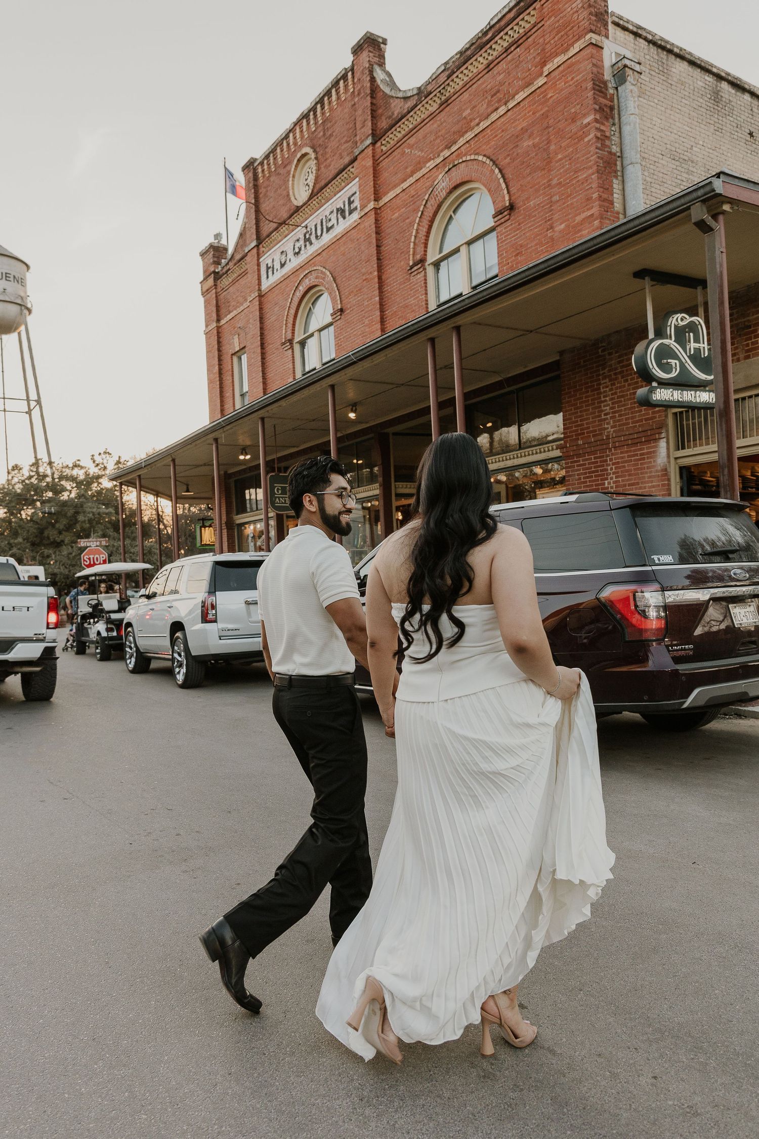 A couple in wedding attire strolls along a brick storefront street with vintage architecture and parked cars.