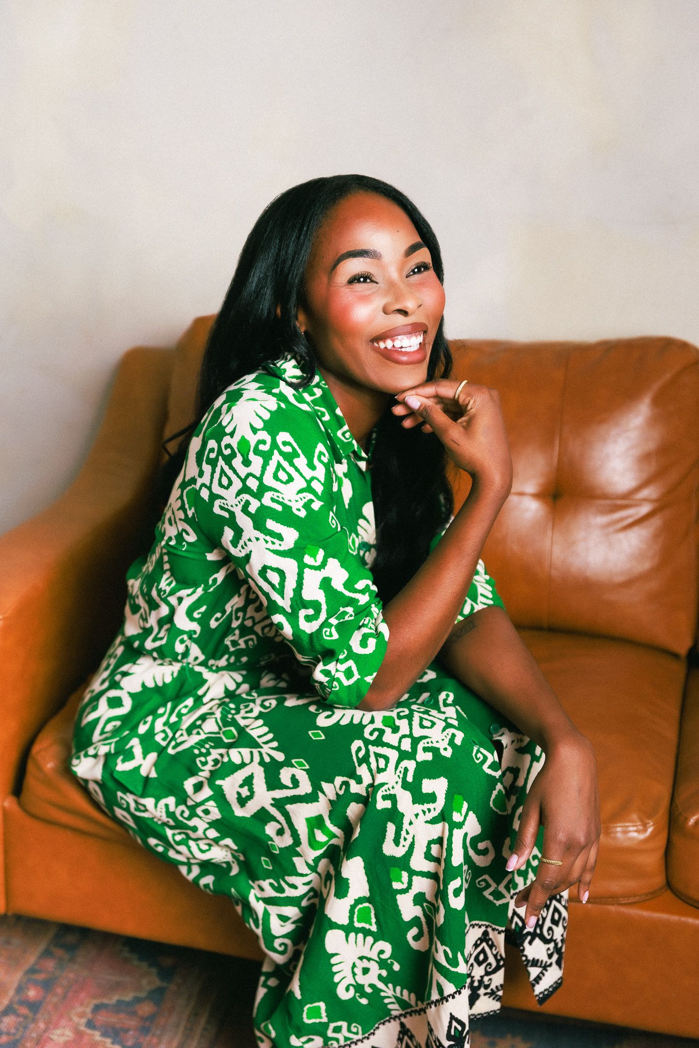 Smiling woman in green patterned dress sitting on brown leather sofa, posing with hand near chin.