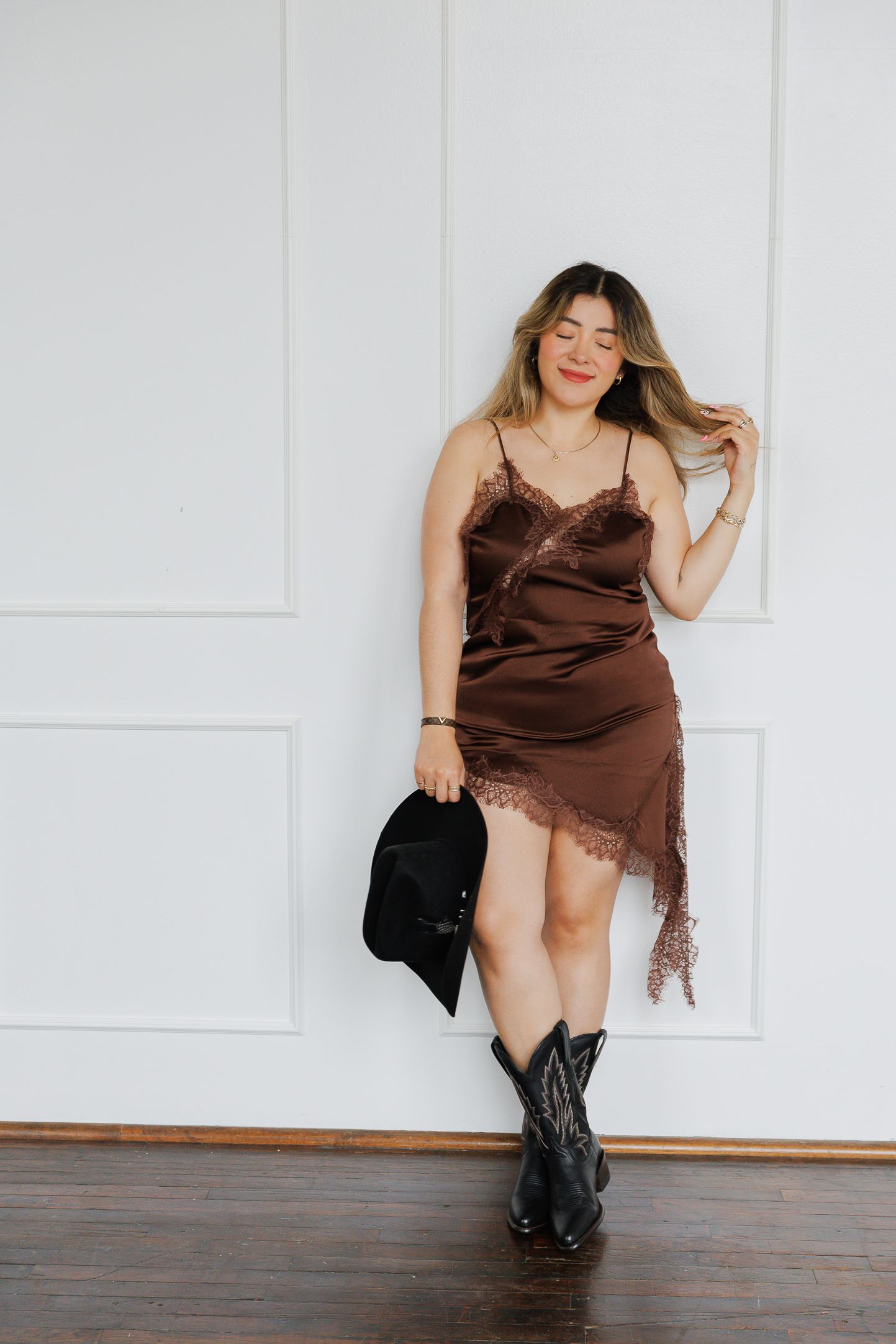 Woman in brown satin slip dress with lace trim, black cowboy boots, holding a hat against white wall.