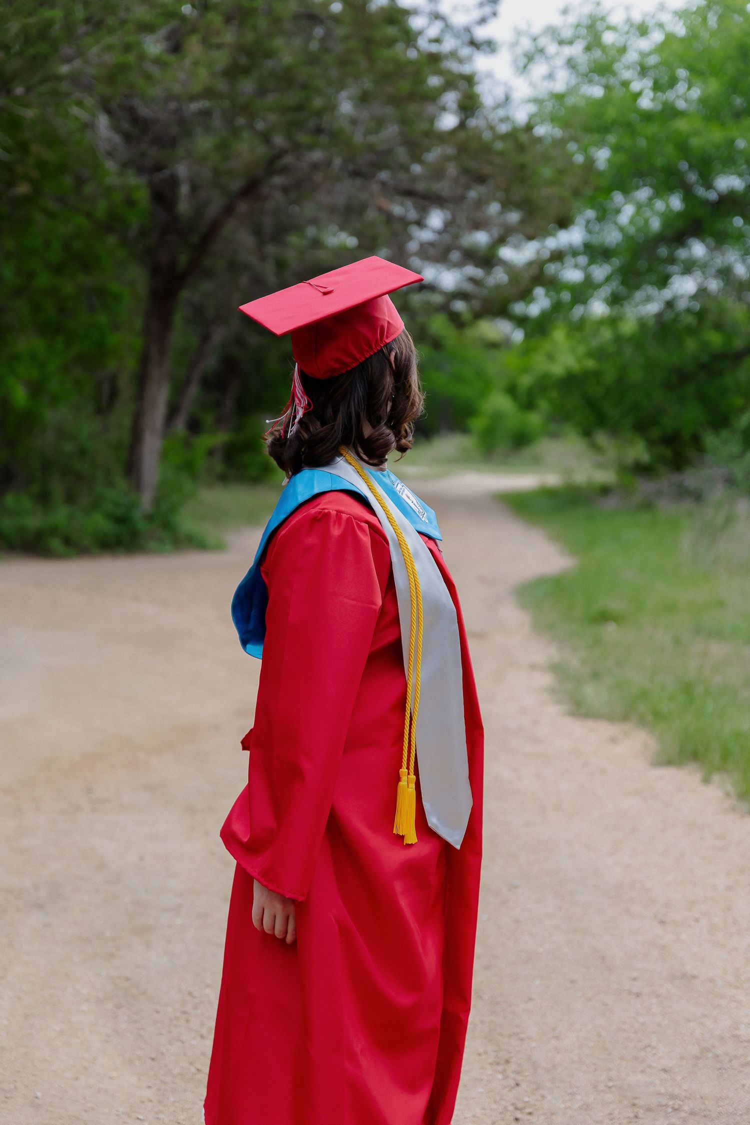 Graduate in red cap and gown looking away on tree-lined path.