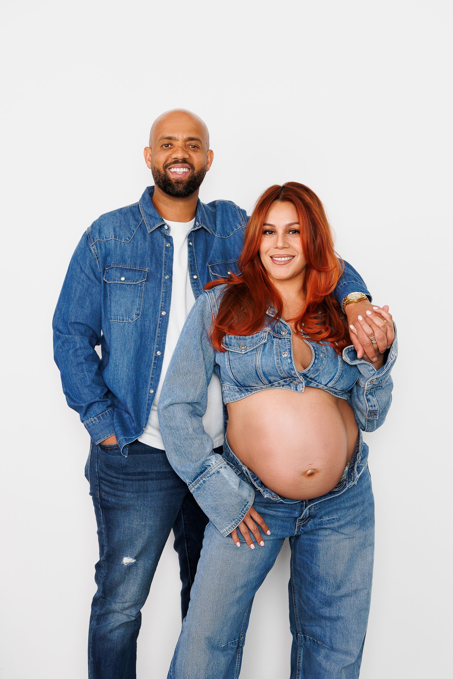 A couple wearing matching denim outfits poses together in a maternity photoshoot showing a pregnant belly.