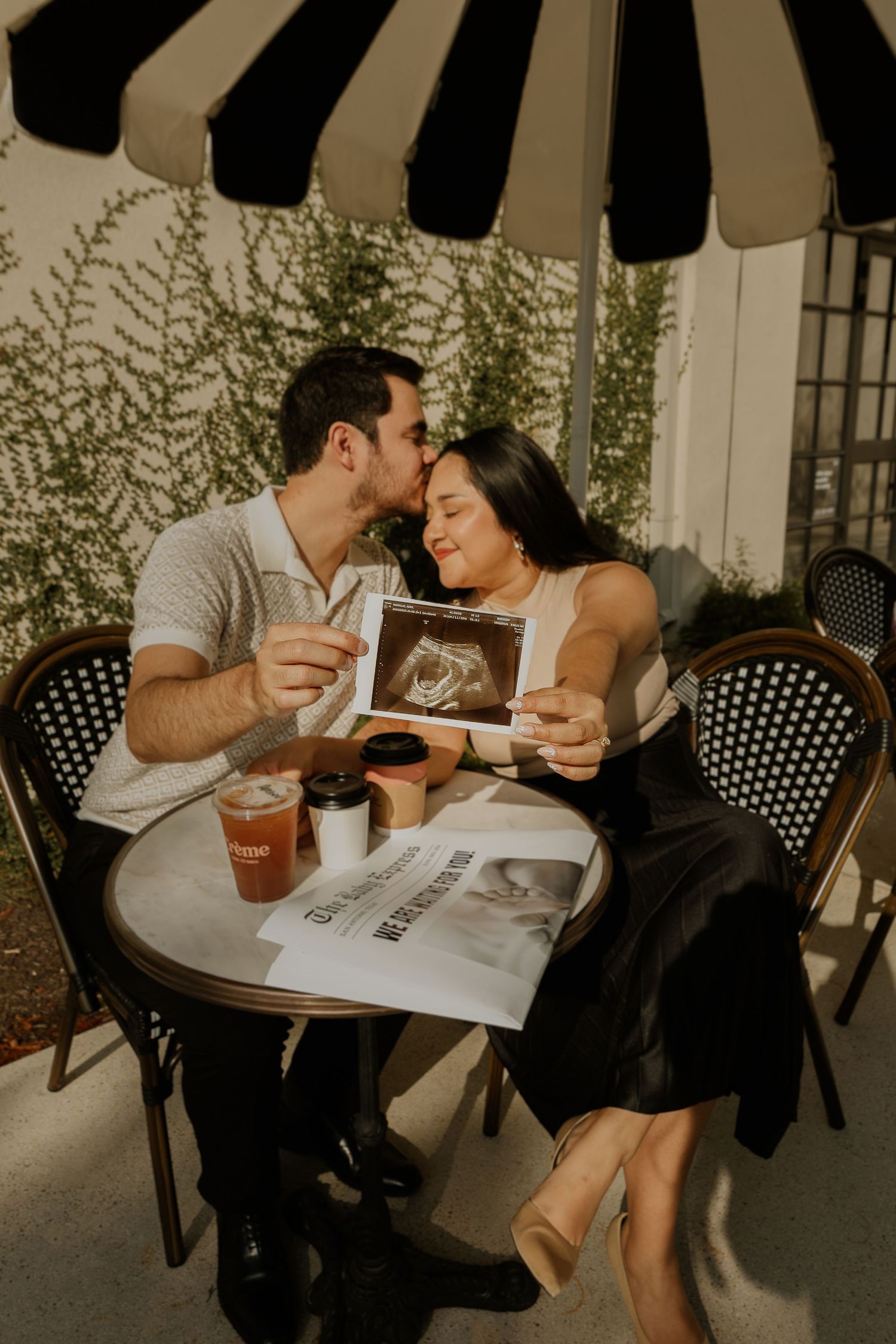 Couple shares intimate moment at outdoor cafe table under striped umbrella with drinks and newspaper.