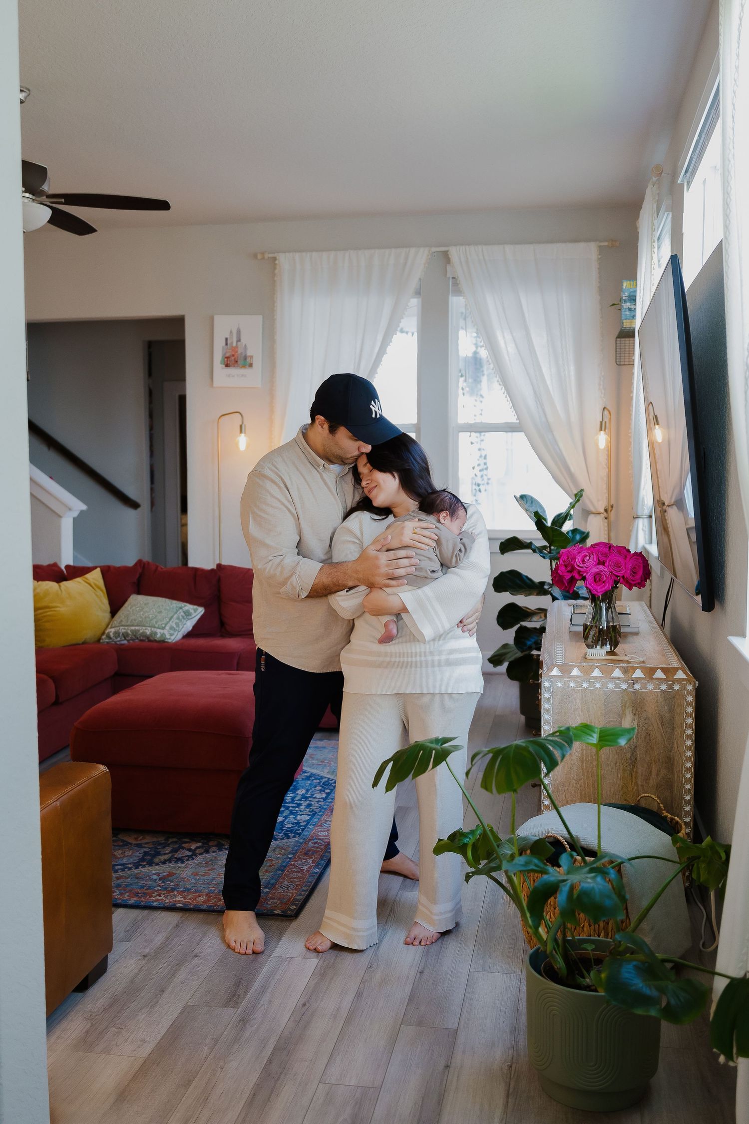 Intimate home portrait of a couple embracing in a cozy living room with plants and sheer curtains.