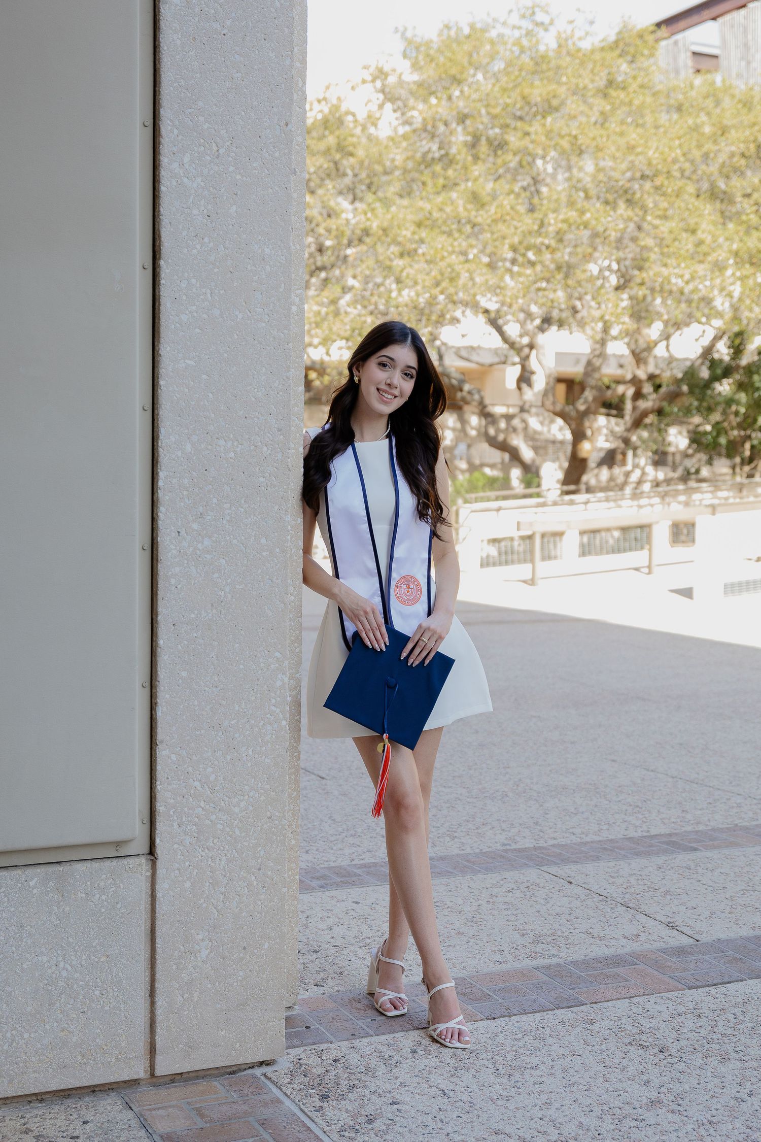 Graduate in white and blue dress poses elegantly against a concrete pillar on campus.