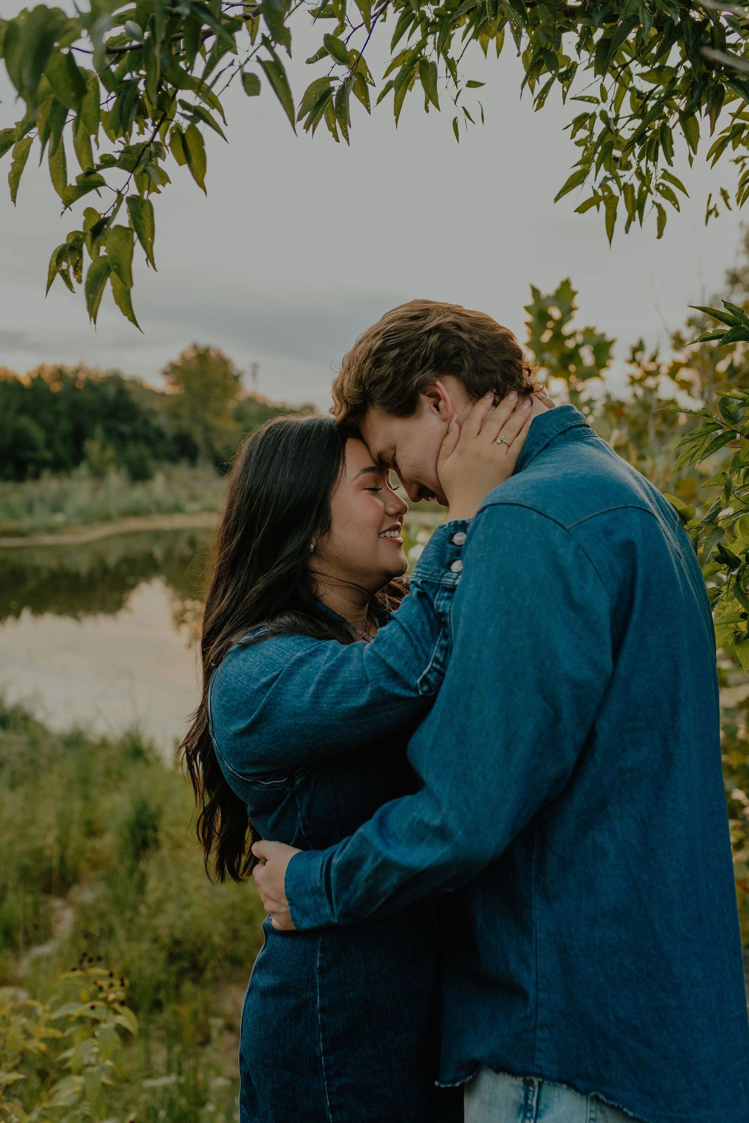 Couple embracing tenderly among trees near a peaceful lake at sunset.