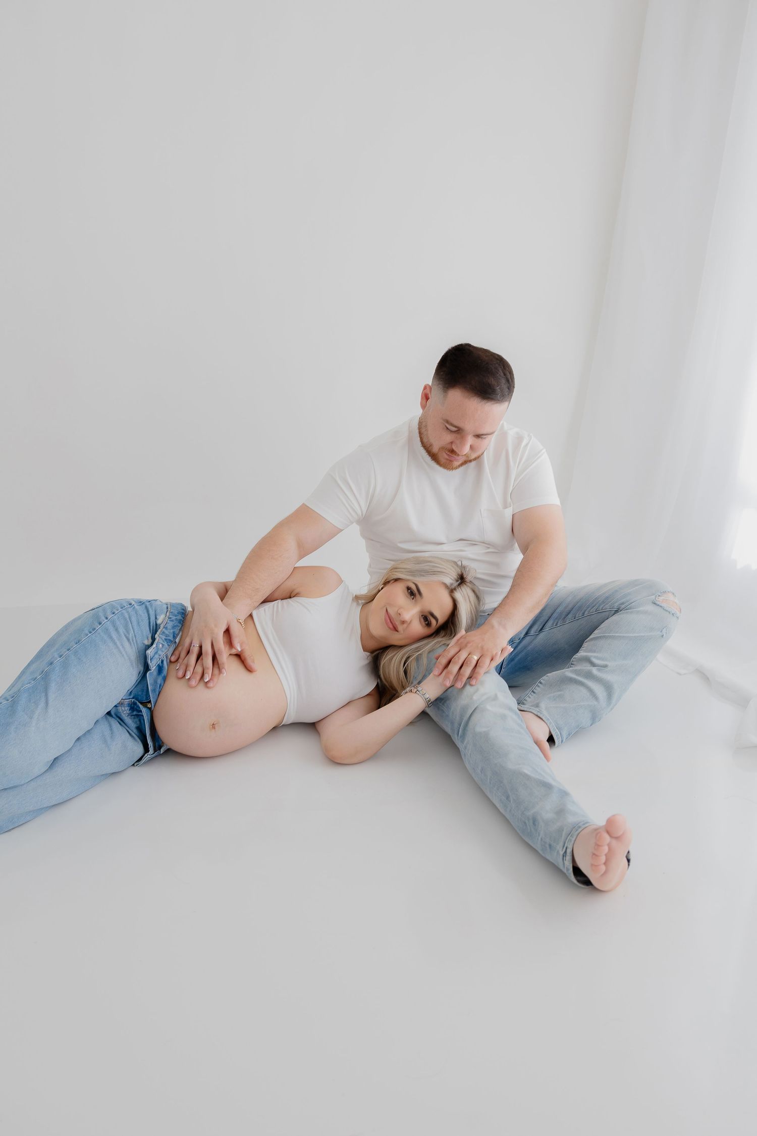 Maternity photo of a couple lying on white floor in casual clothing.