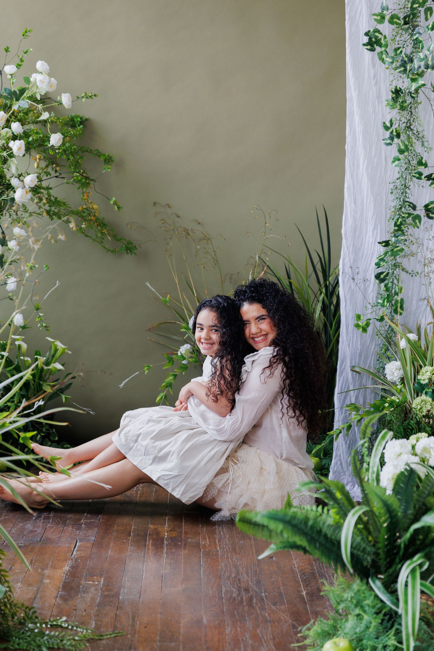 Two people in white dresses lay on wooden floor with green plants and white curtains creating dreamy atmosphere.