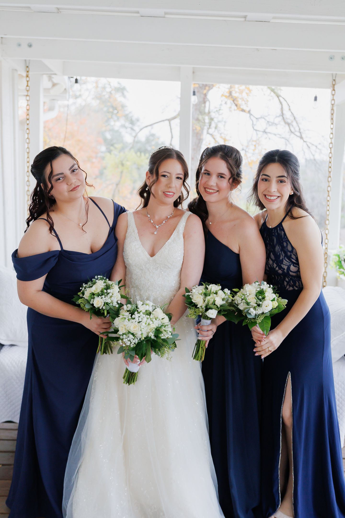 Wedding party in navy blue dresses and white gown laughing together in sunlit room with white bouquets.