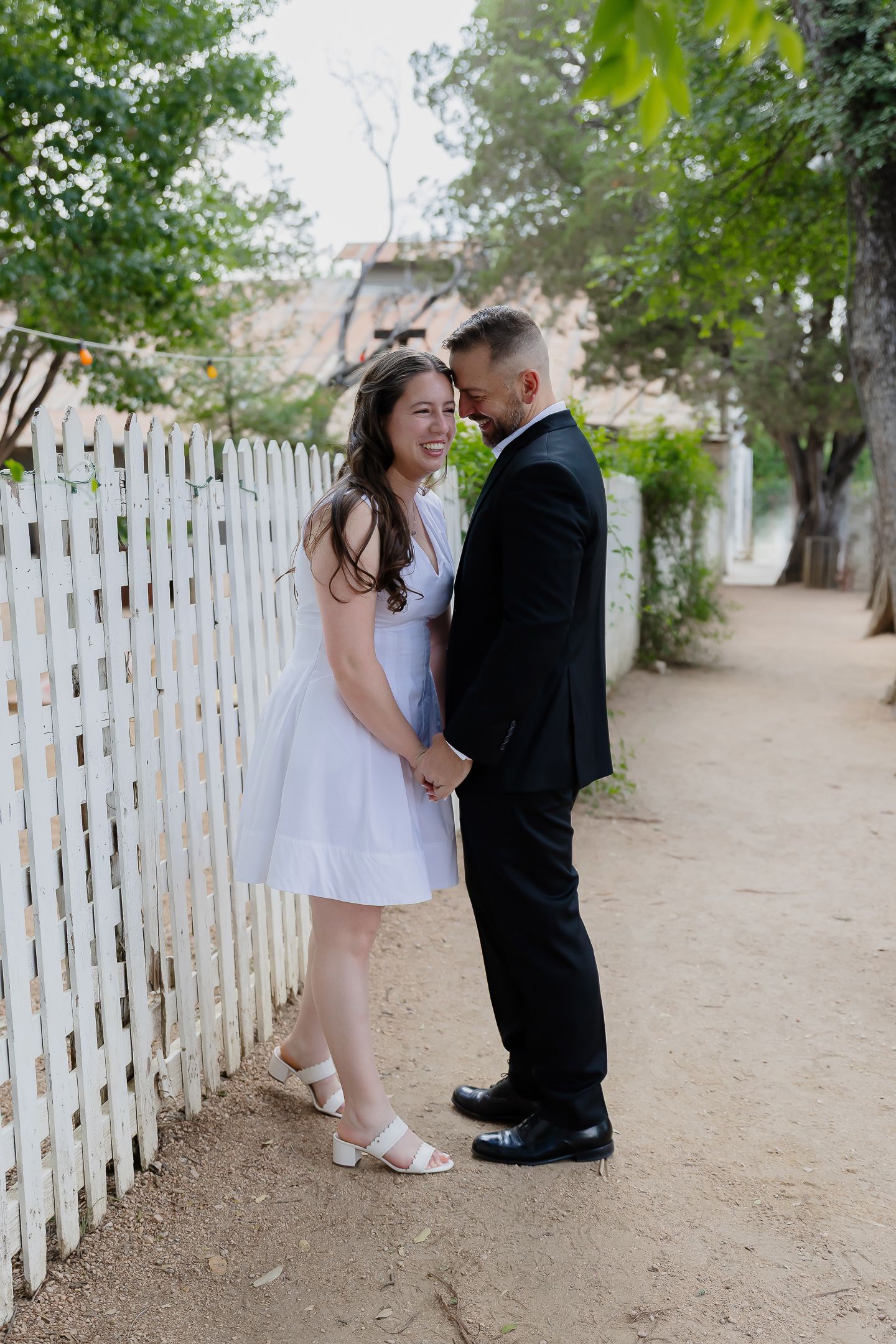 A couple shares a romantic moment along a white picket fence on a dirt path surrounded by greenery.