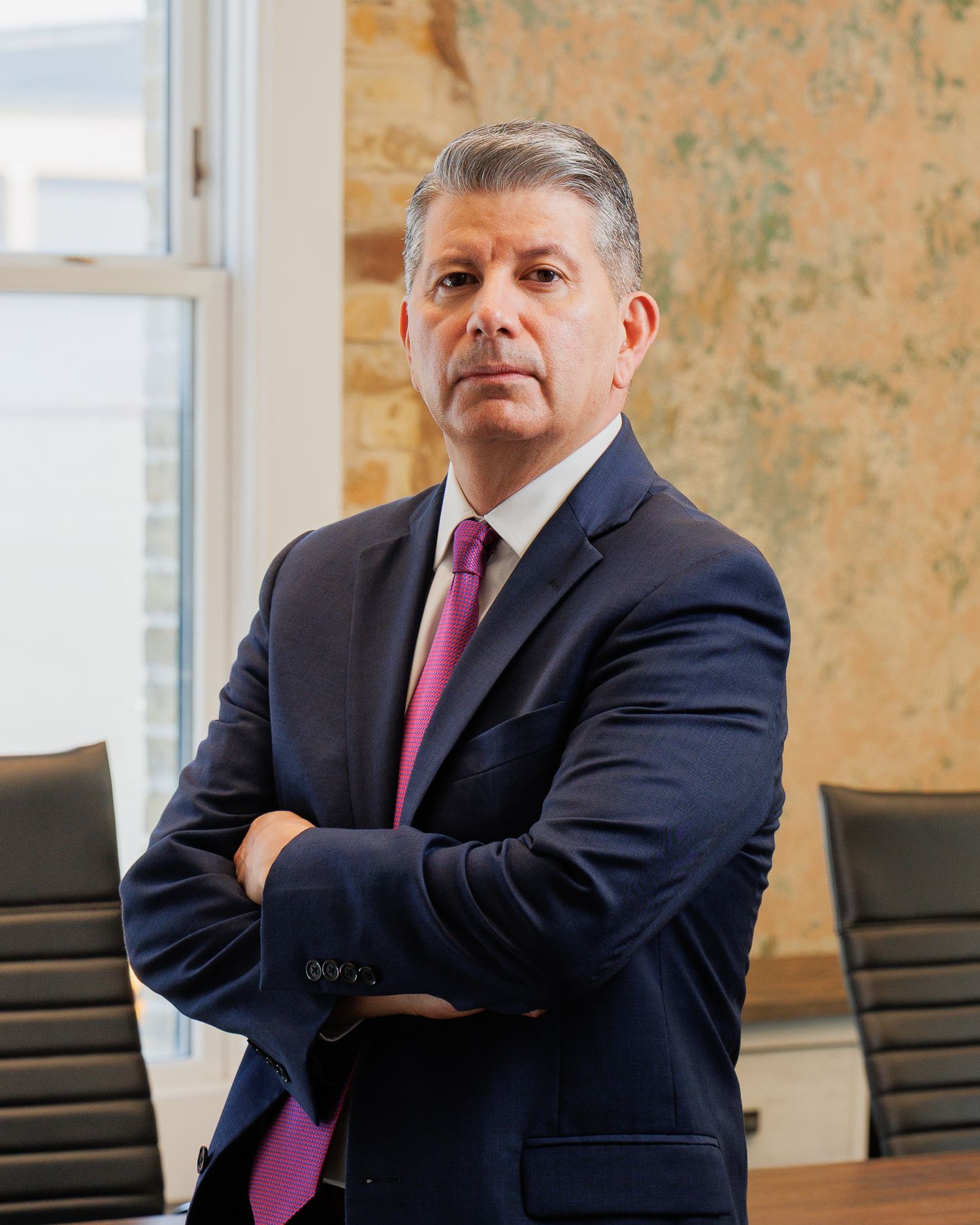 Professional attorney in navy suit and pink tie stands with arms crossed in a modern conference room.