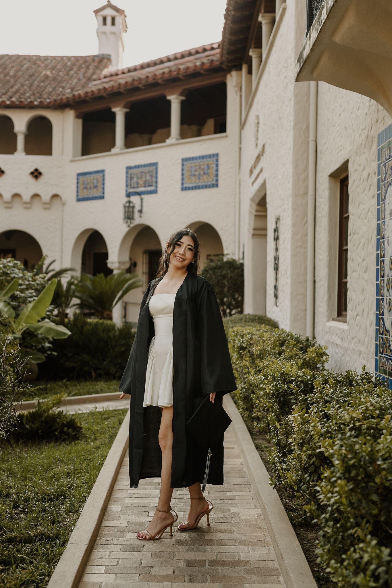 Graduate in black gown posing along Mediterranean-style courtyard walkway.