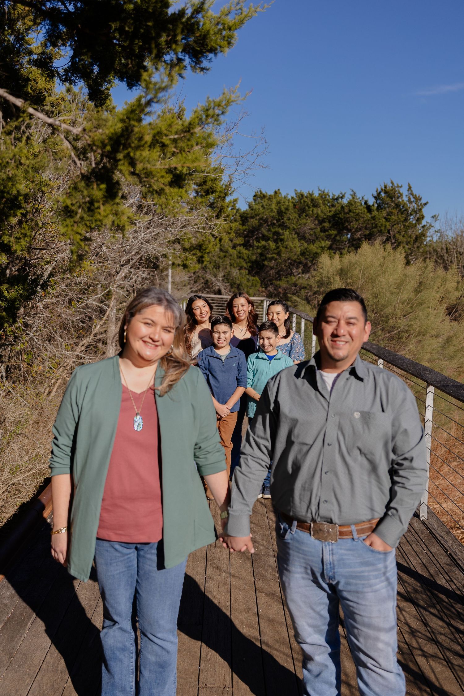 A group of people stand together on a wooden boardwalk trail surrounded by evergreen trees on a sunny day.