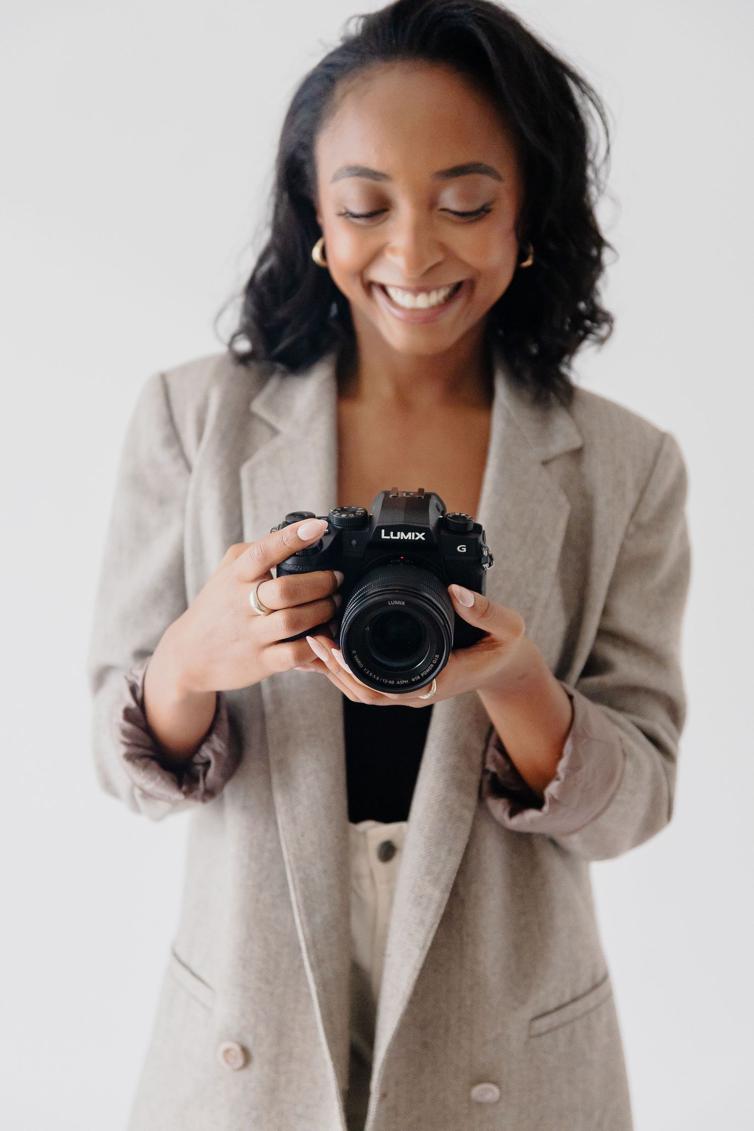 Professional photographer in beige blazer smiling while holding a Lumix camera.