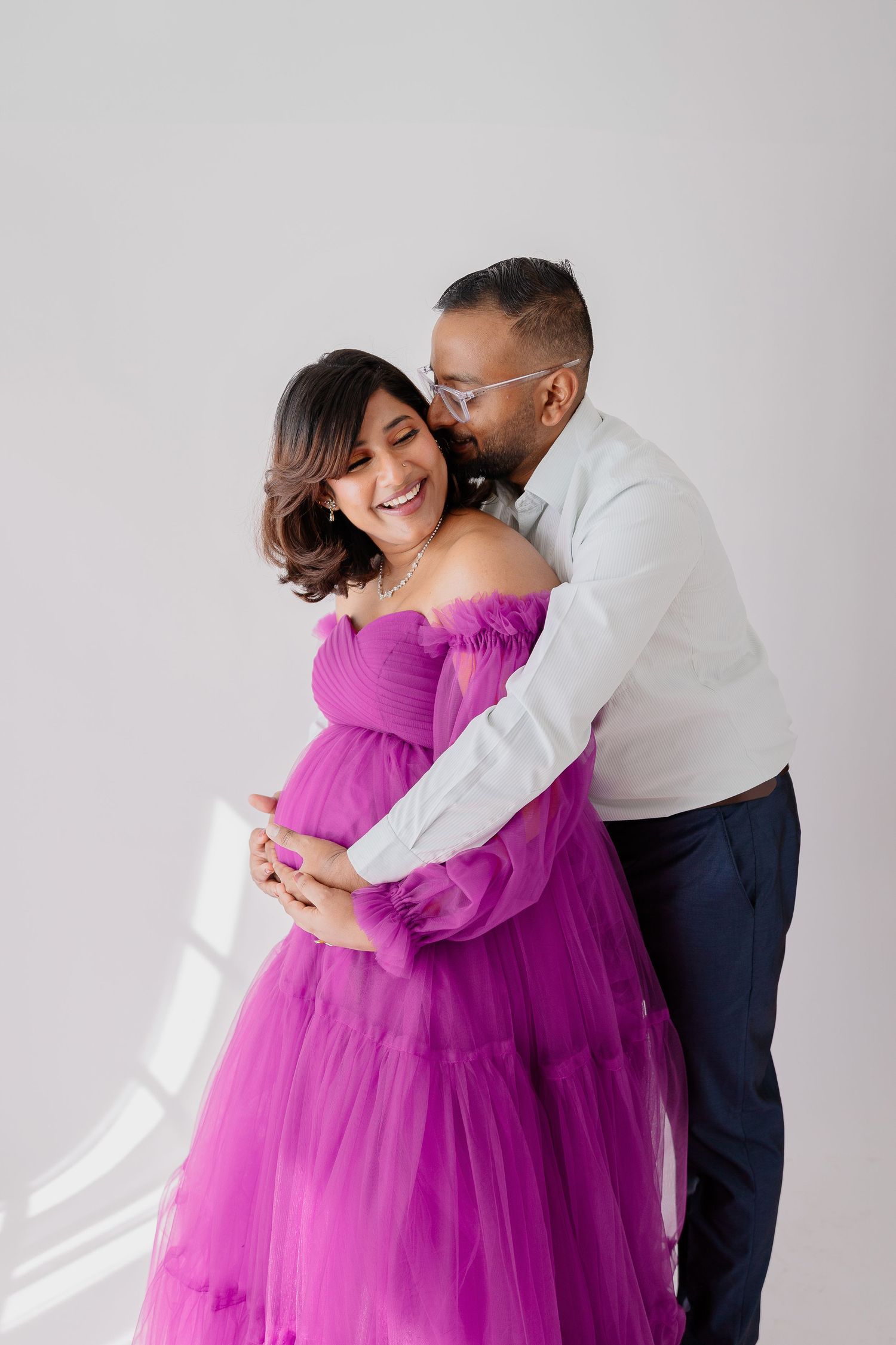 A couple shares an intimate moment in a maternity shoot, with the woman wearing a flowing purple gown.