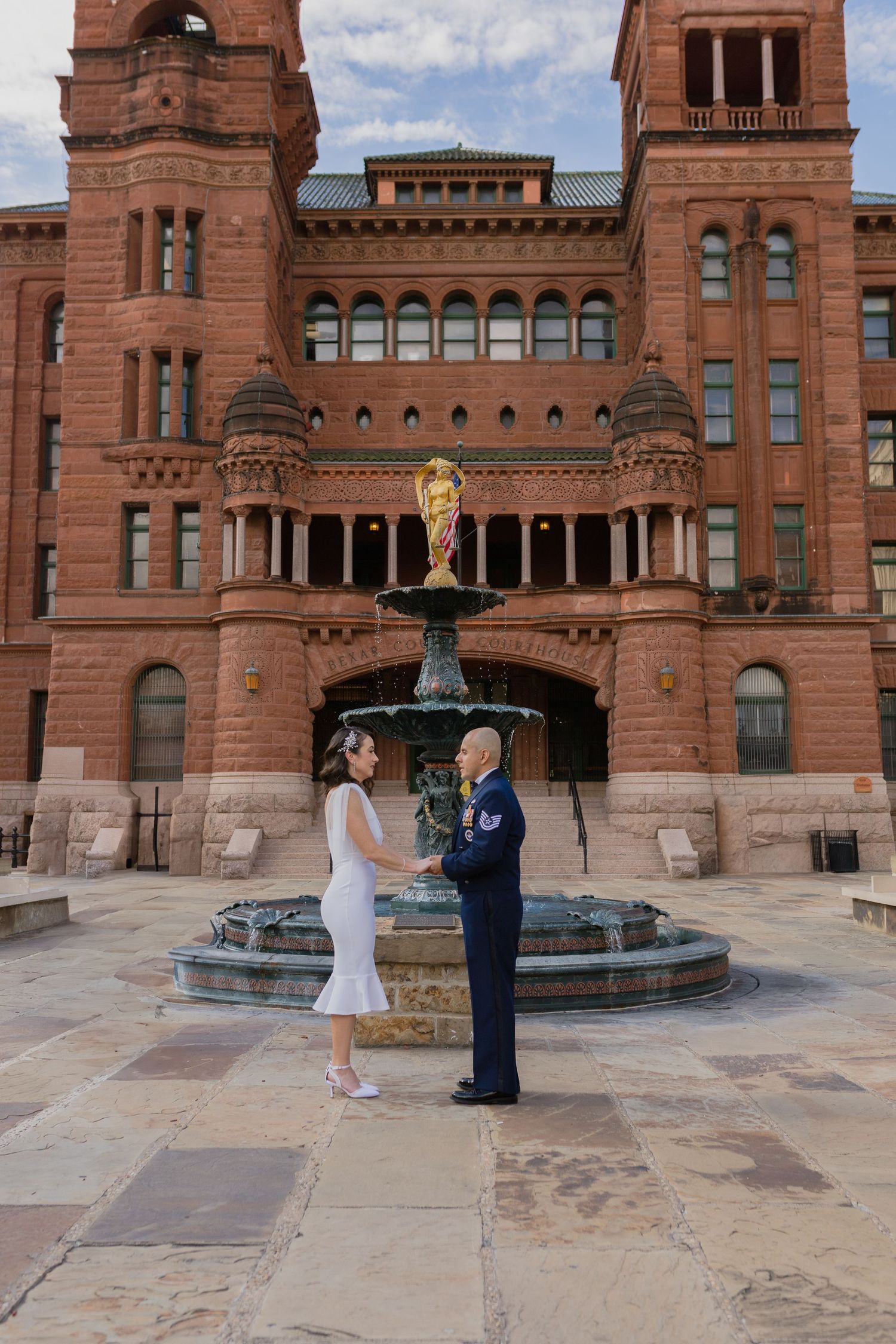 A couple exchanges vows in front of a grand historic courthouse with ornate architecture and fountain.