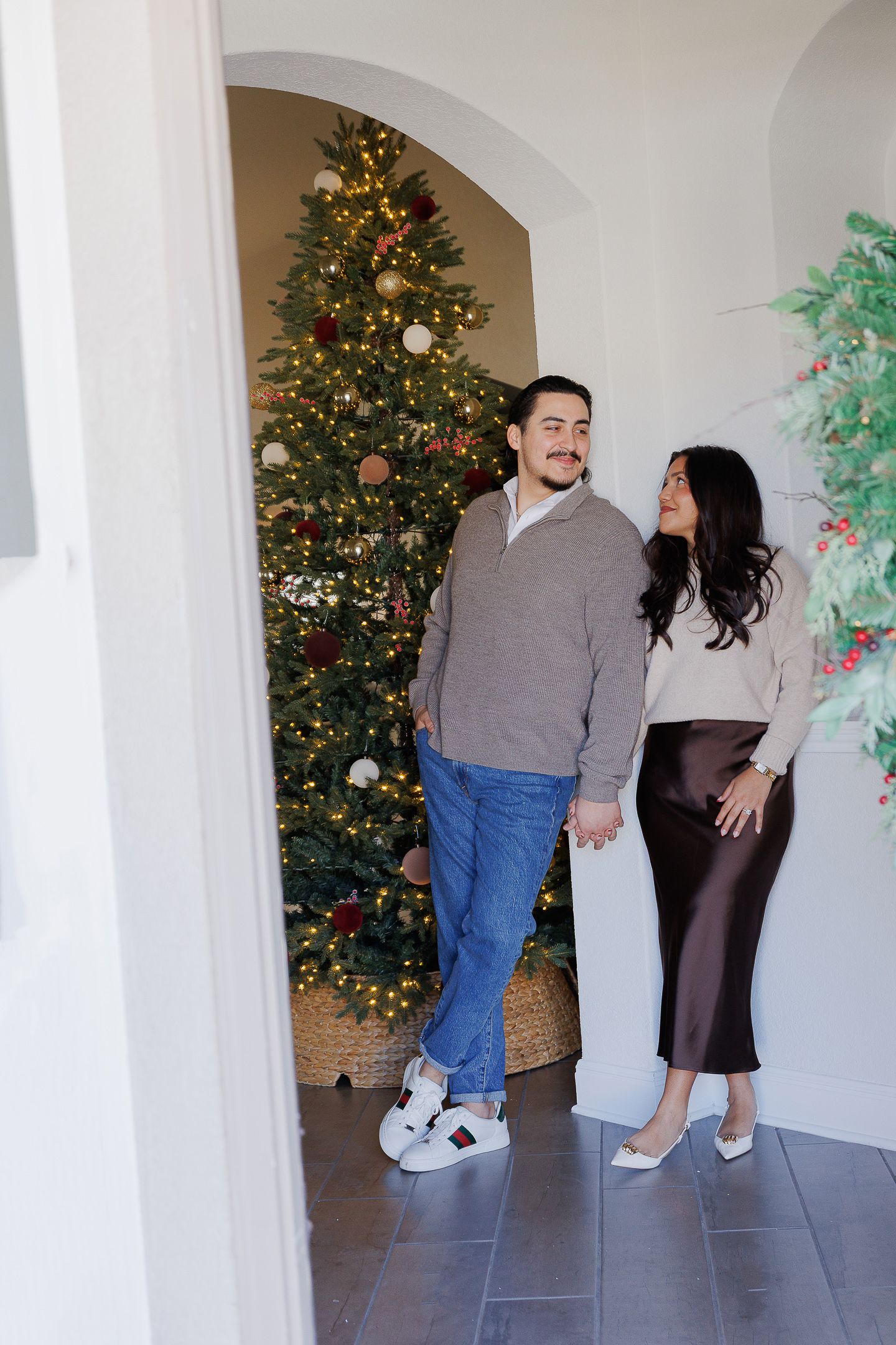 A couple poses together in front of a decorated Christmas tree in a bright interior space with white columns.