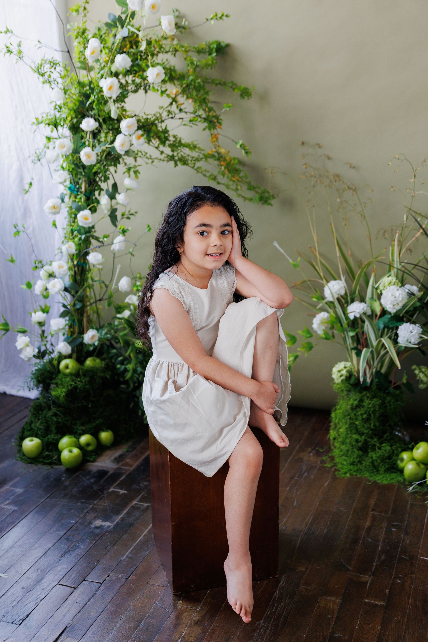 A child sits on a wooden floor against a wall decorated with lush green vines and white floral arrangements.
