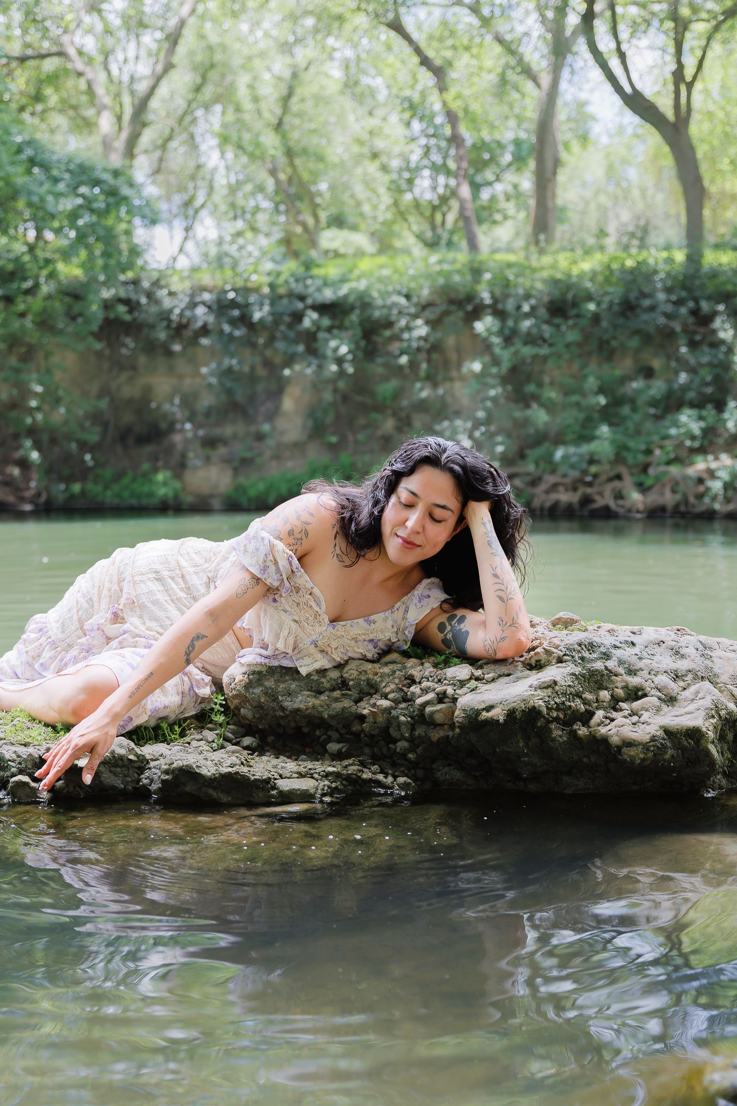 A woman in a floral dress lies on mossy rocks by a serene pond in a lush green garden.