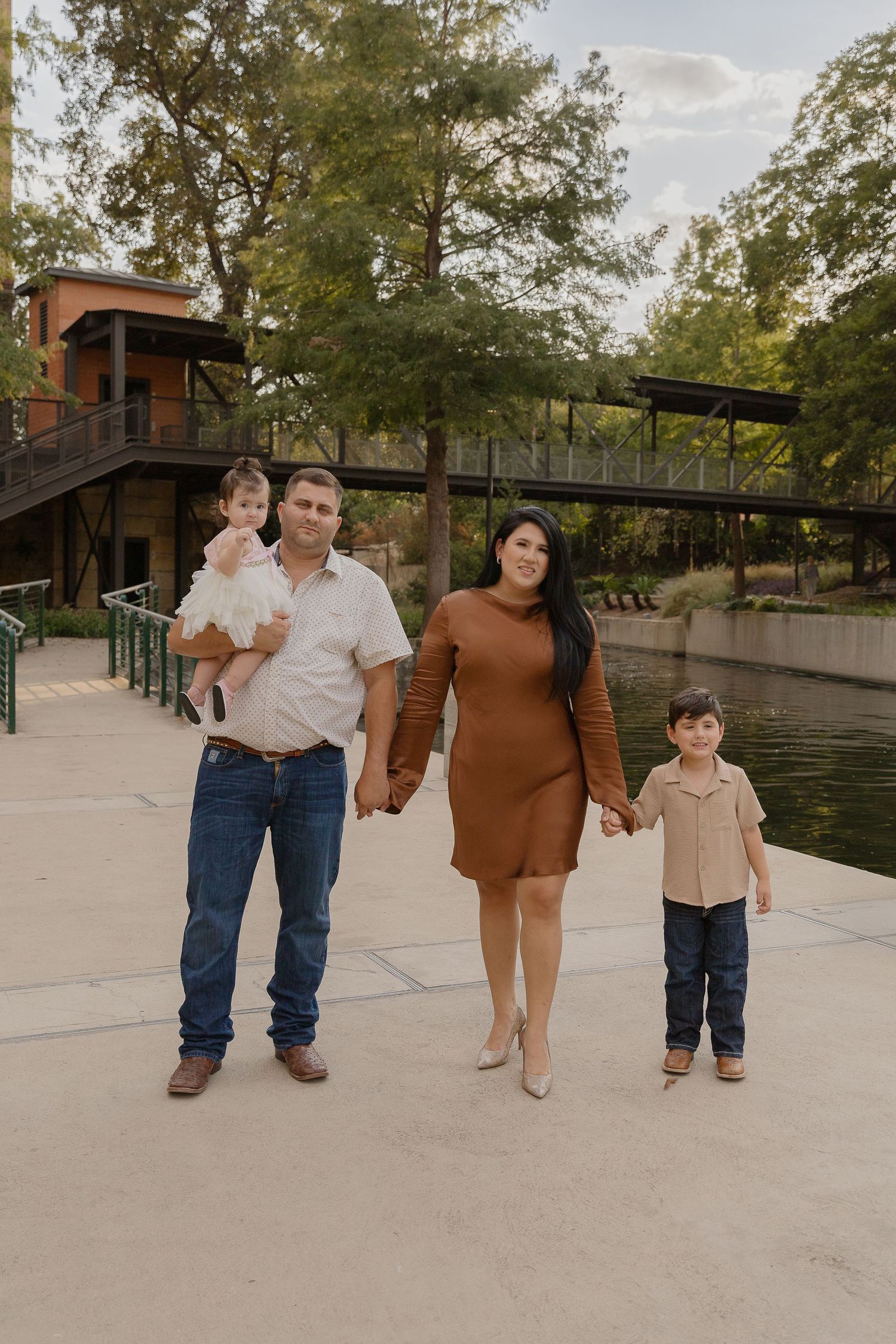 A family stands together on a path near a waterfront boardwalk and treehouse on a sunny afternoon.