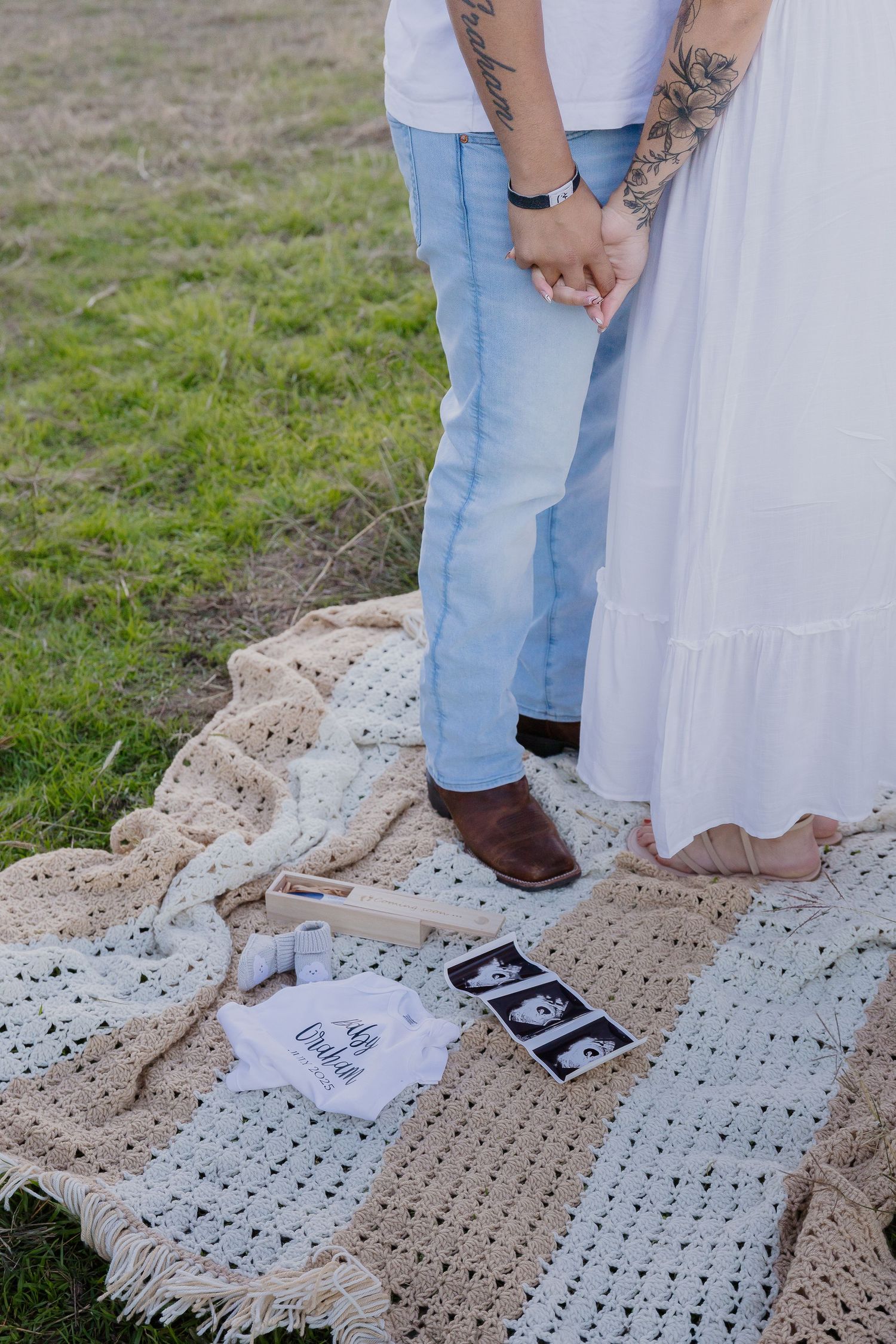 Couple holding hands on a picnic blanket with ultrasound photos laying nearby on the grass.