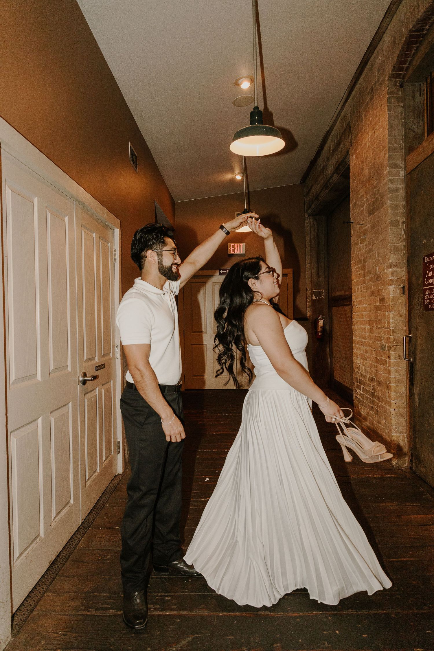 A couple dances playfully in a rustic hallway with brick walls and hardwood floors.