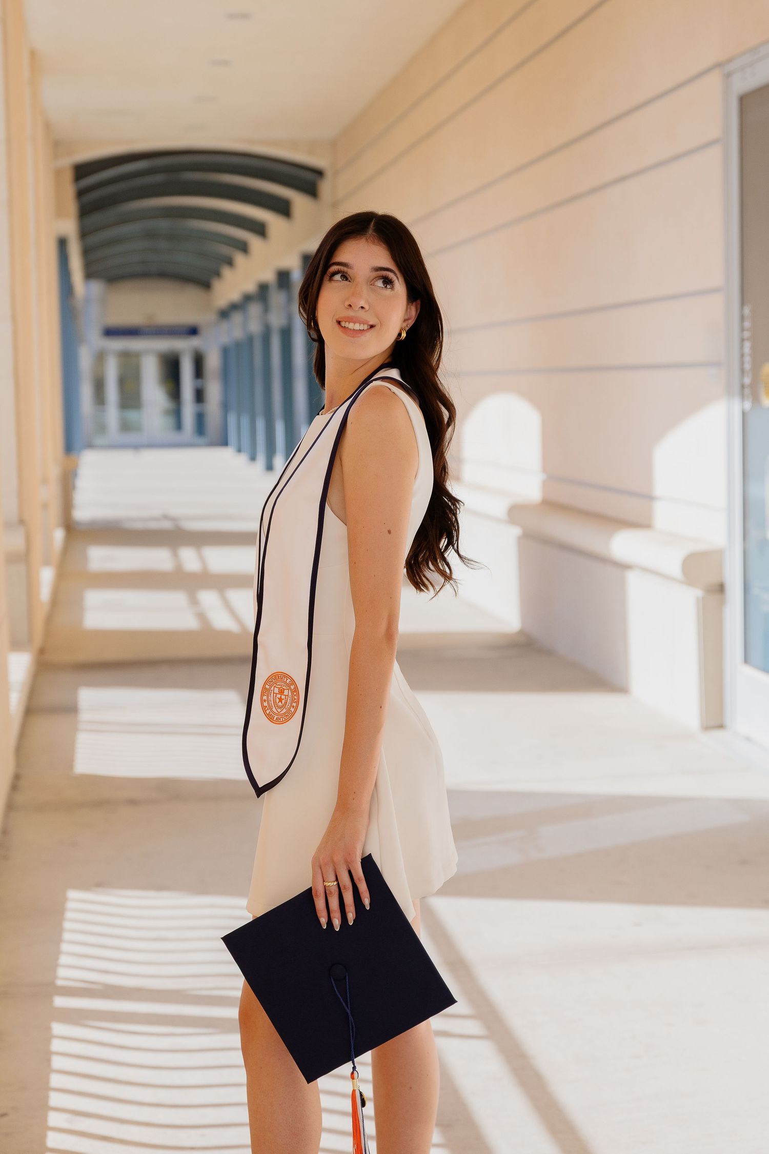 Graduate in white dress and graduation stole poses in sunlit campus colonnade.