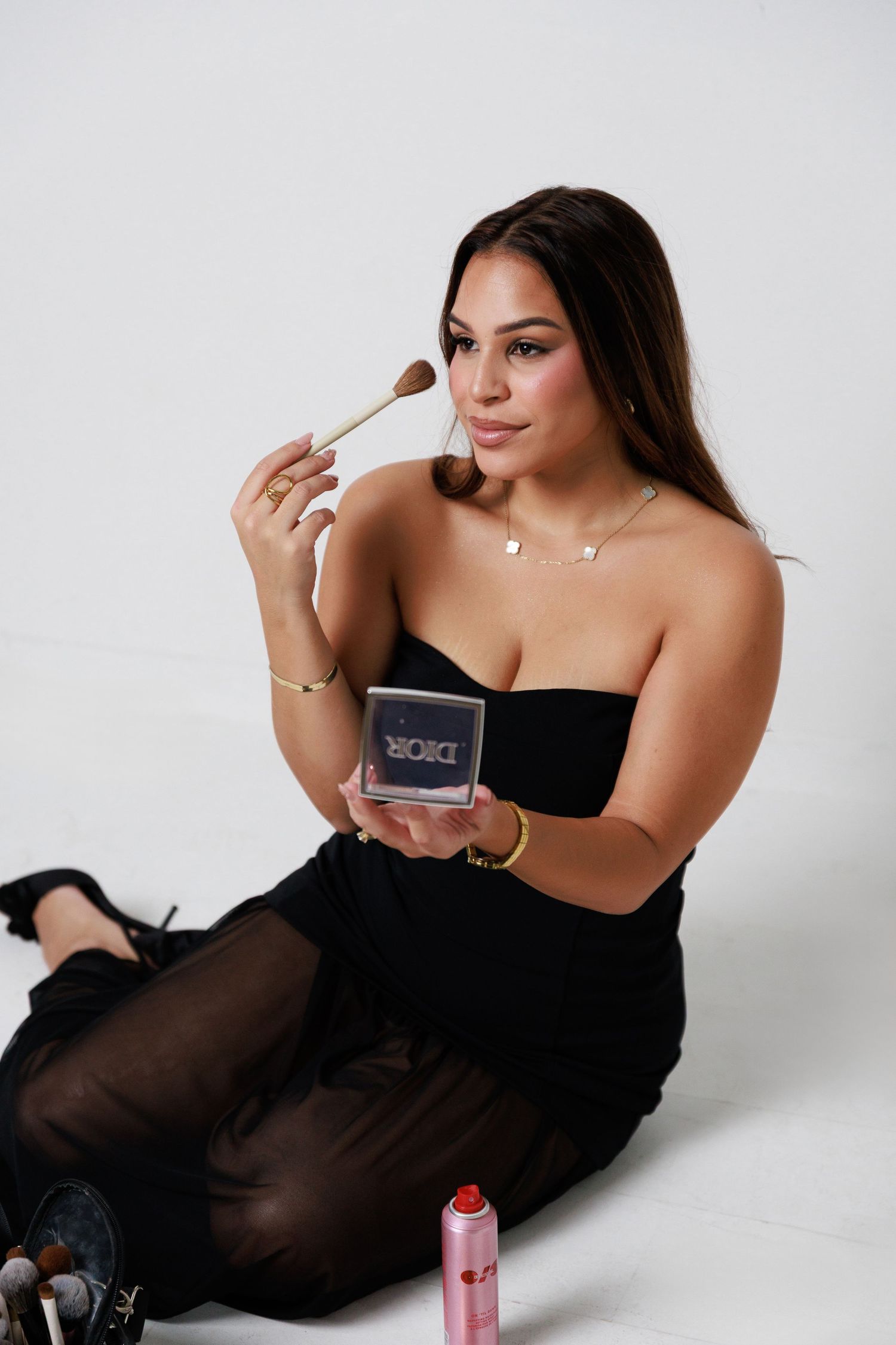 A person in a black dress poses on a white backdrop while holding makeup brushes and cosmetics.
