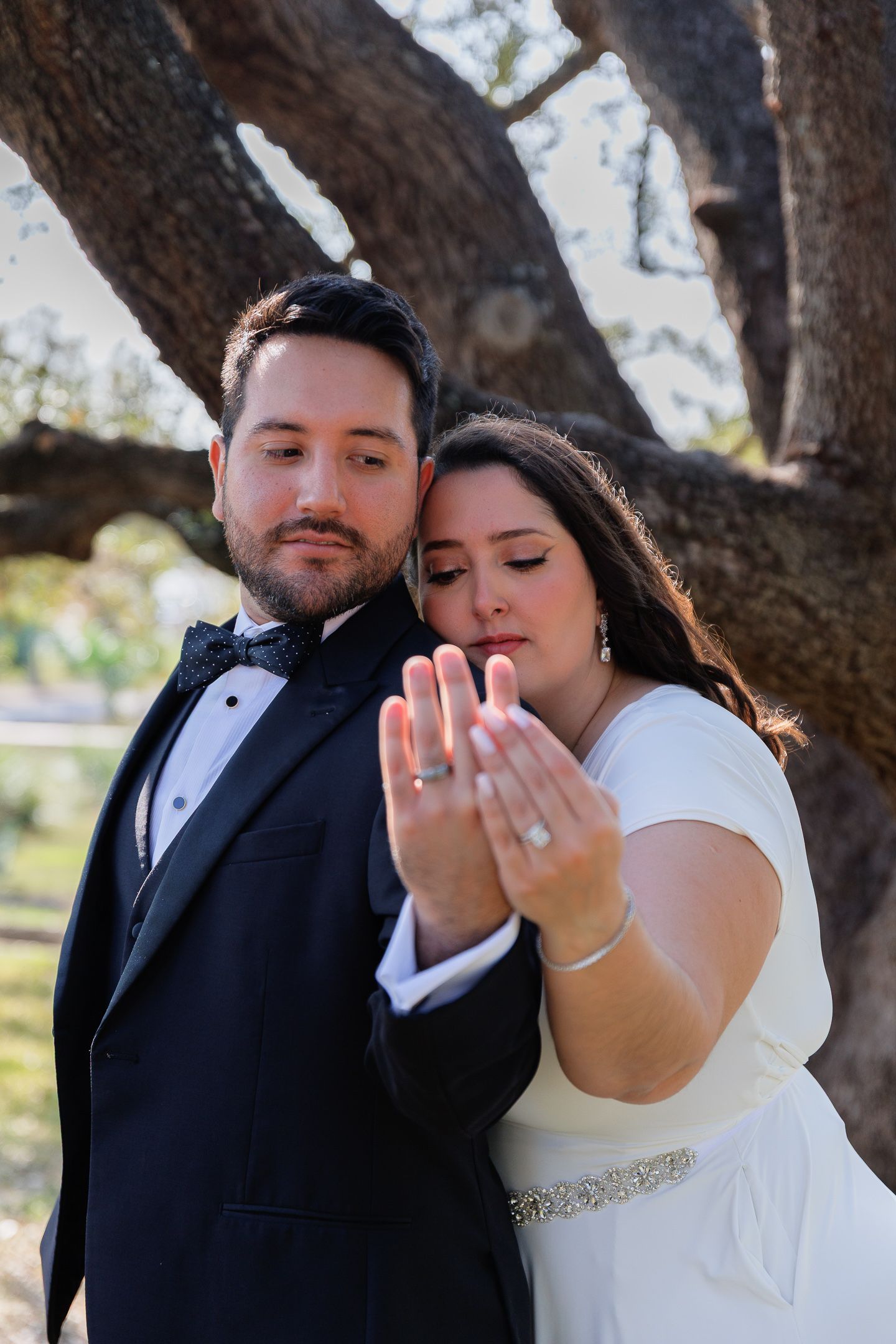 Romantic wedding couple poses together under tree branches in formal attire during outdoor photo session.