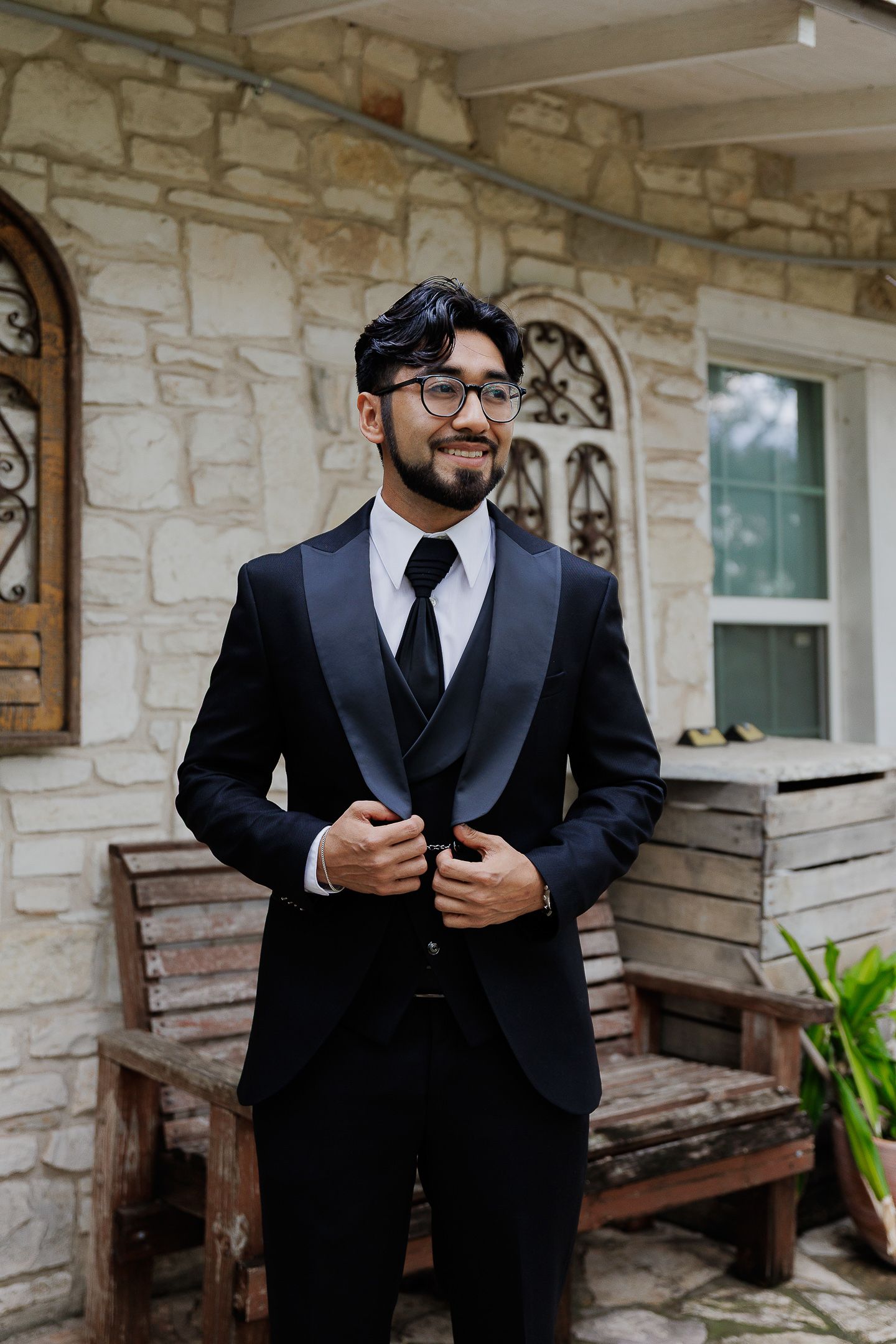 A person in a formal black tuxedo and tie stands on a stone porch with wooden benches in the background.