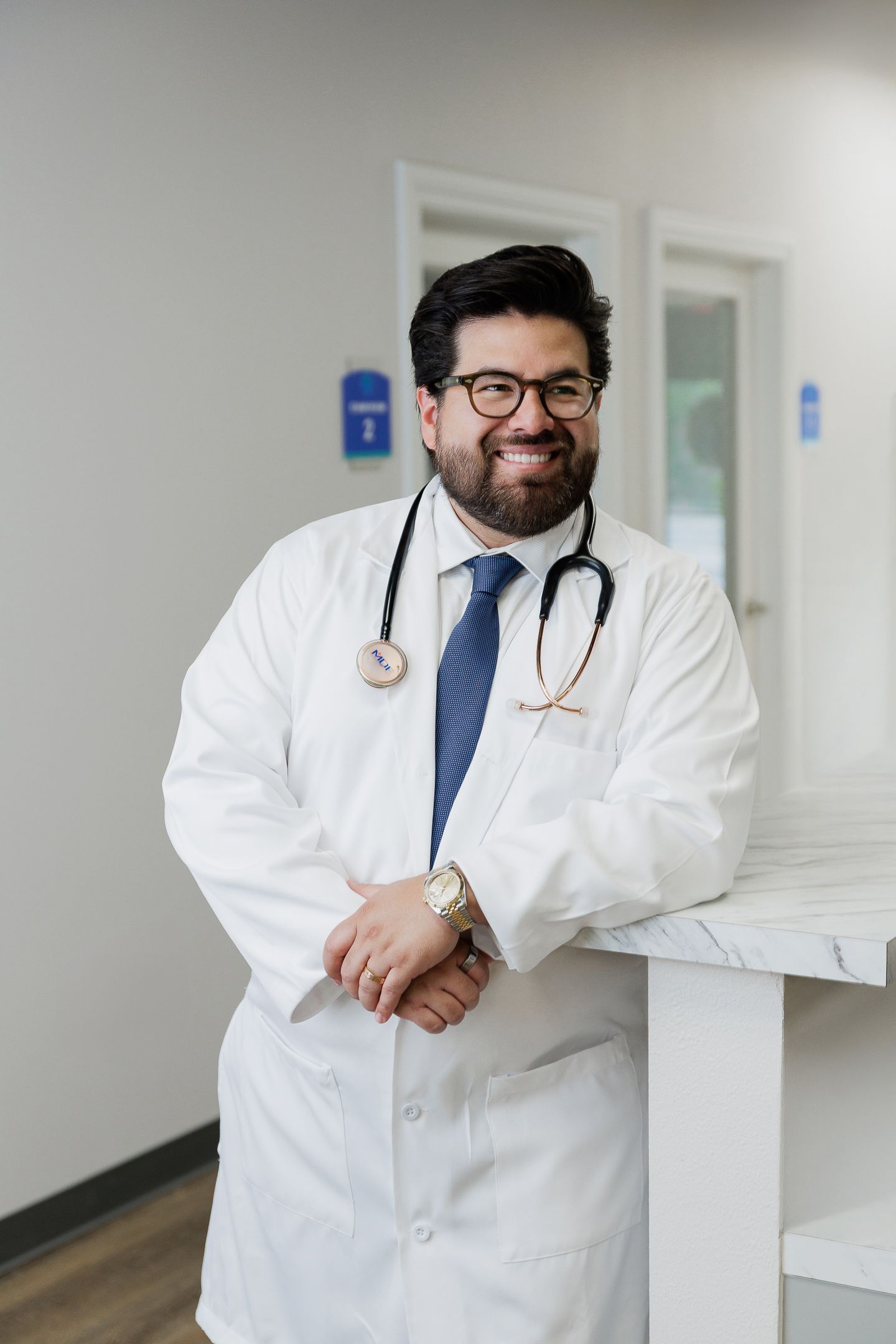 Medical professional in white coat and stethoscope smiling while leaning against wall in hospital hallway.